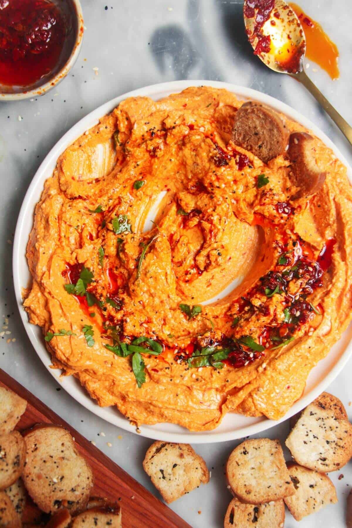 Spicy harissa feta dip on a small white plate, with bagel chips and more harissa on the side.
