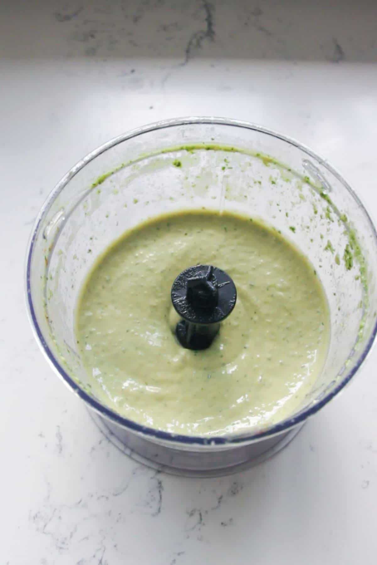 Basil and tahini mixed in a small food chopper on a white marble background.