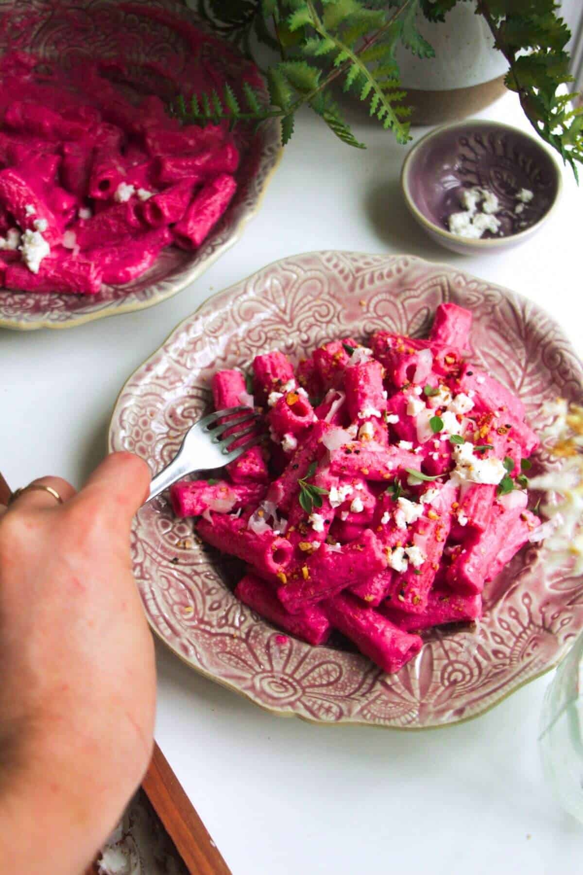Hand holding a fork digging into plate of bright pink pasta.