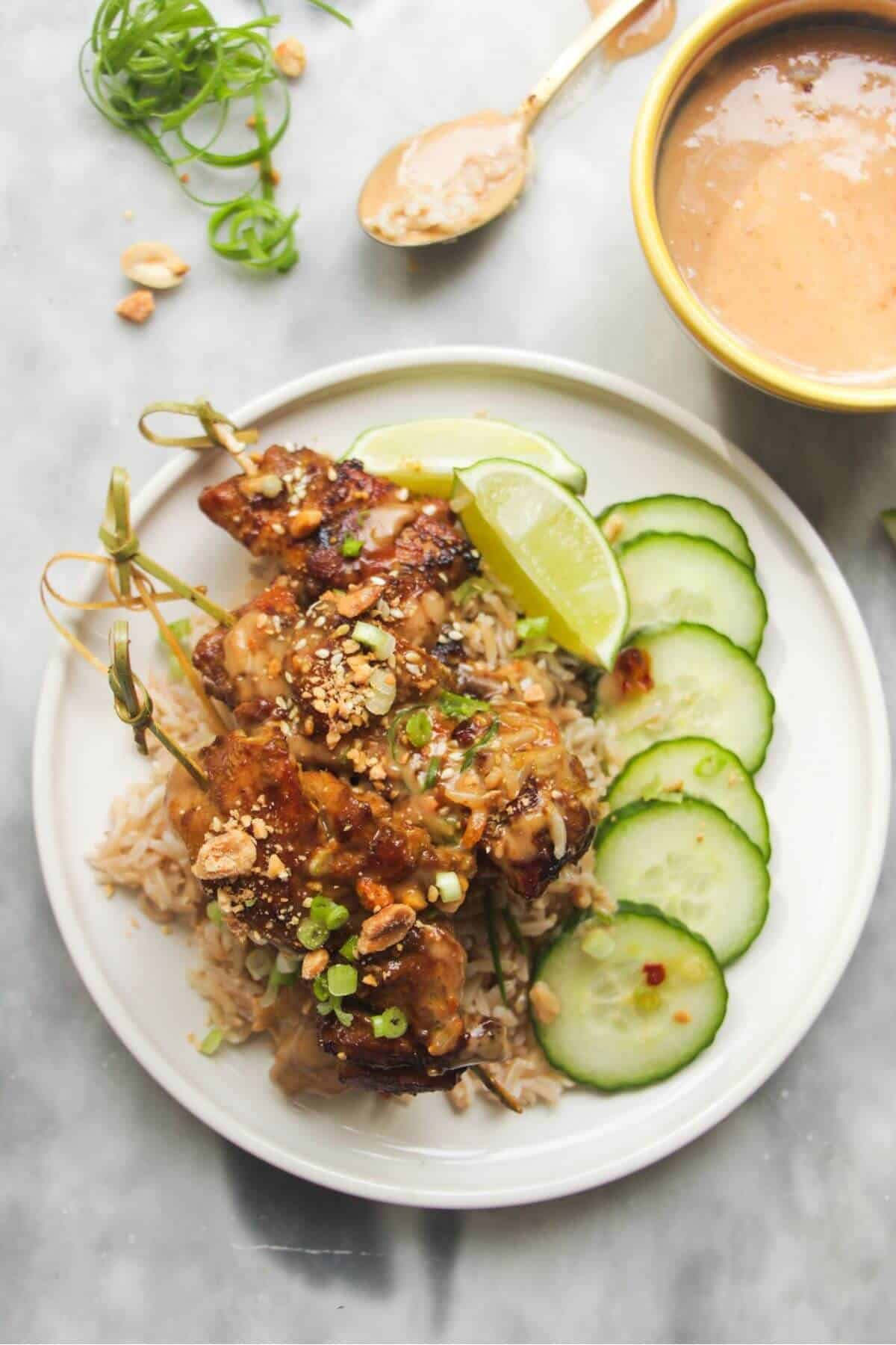 Chicken satay skewers on rice with cucumber slices on the side, with a small bowl of satay peanut sauce behind.