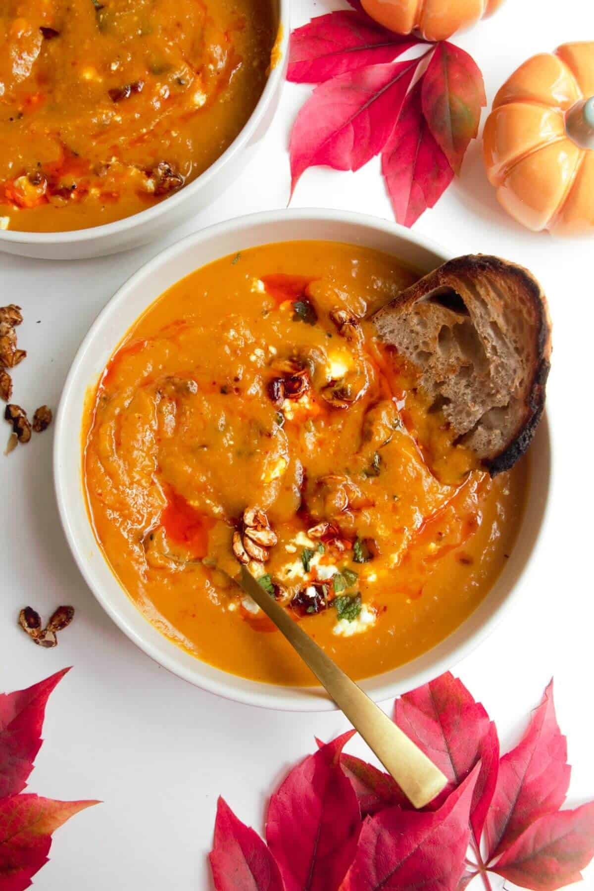 Roasted butternut squash soup in a white bowl with a gold spoon, topped with chilli oil, feta and squash seeds.
