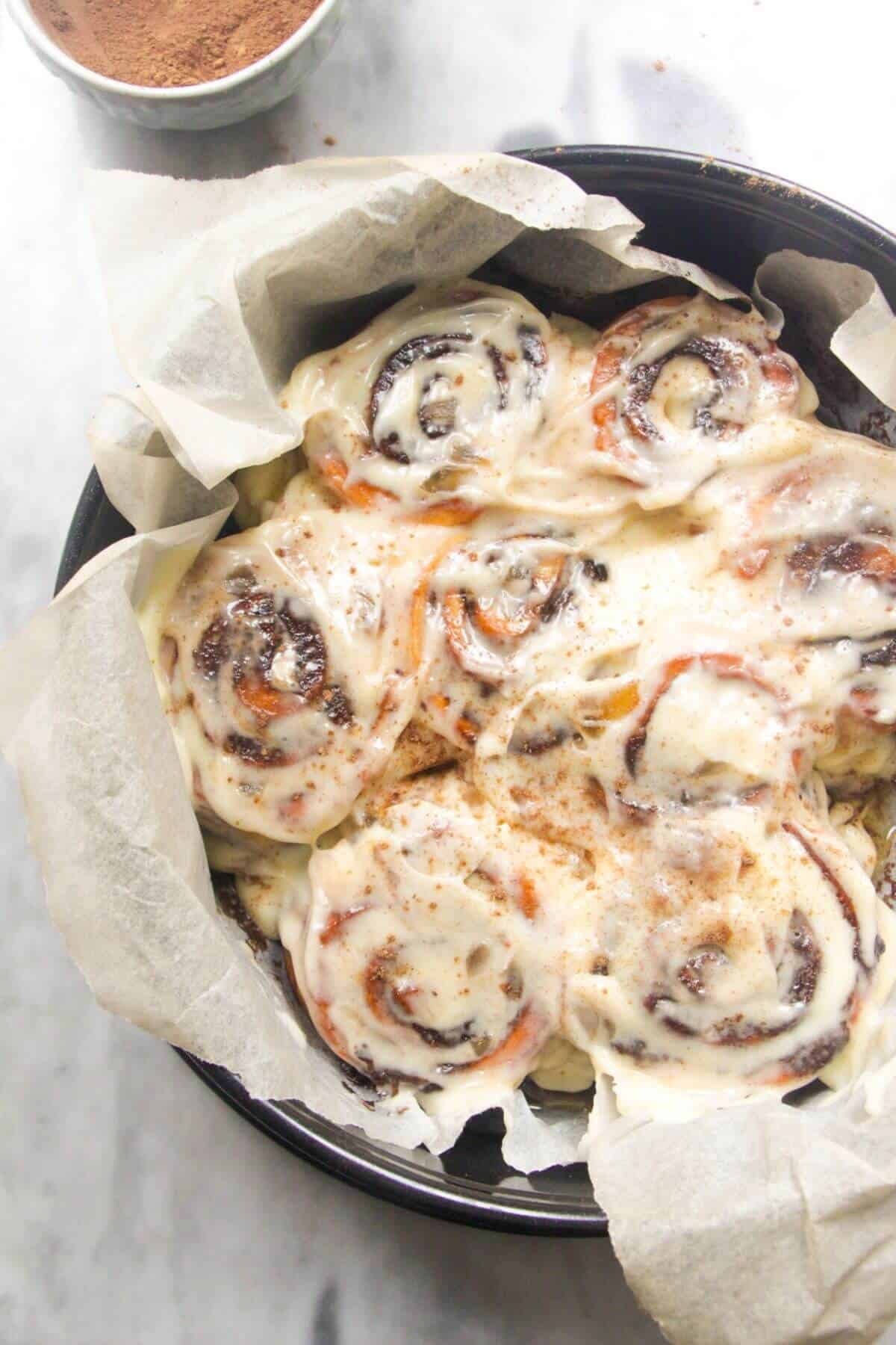 8 baked and iced cinnamon rolls in a baking paper lined cake tin with pumpkin spice in a small bowl in the background.