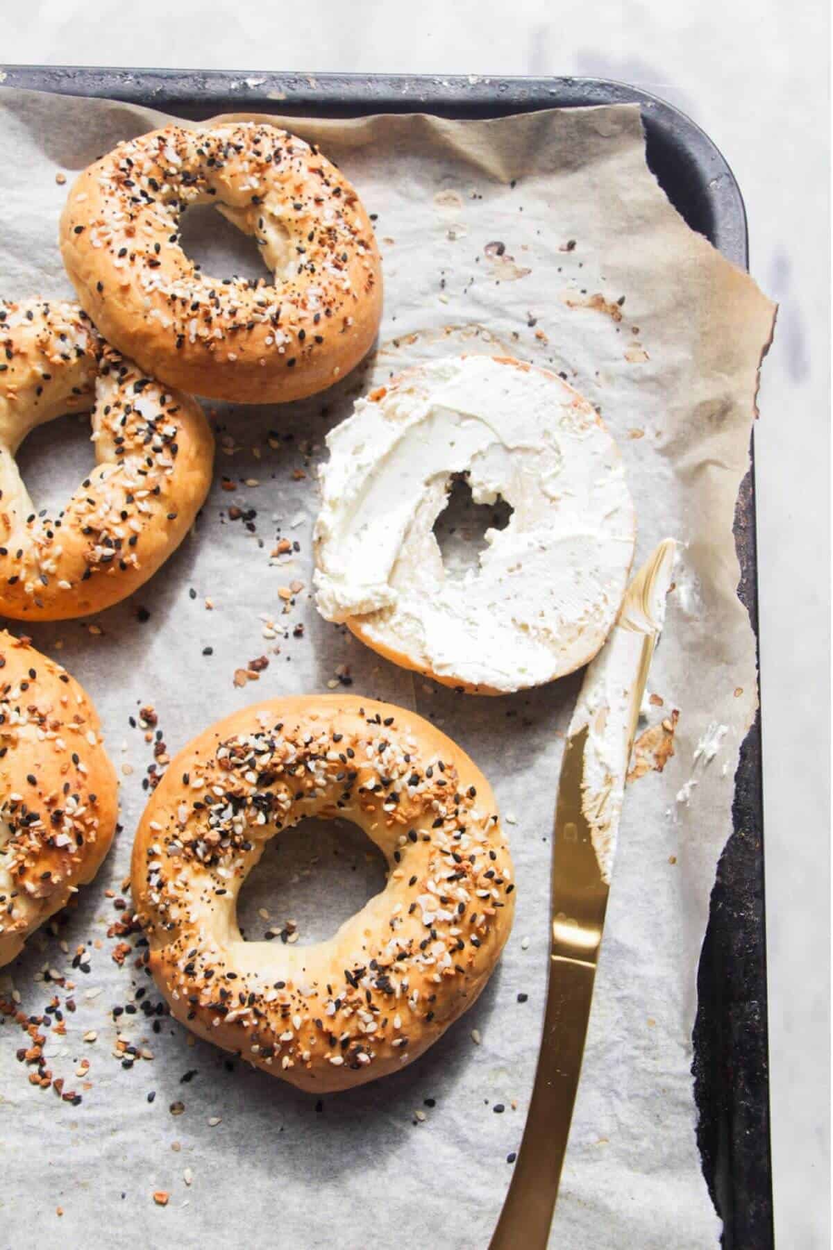 Cut bagel with cream cheese smeared on it on an oven tray, with more bagels on the side.