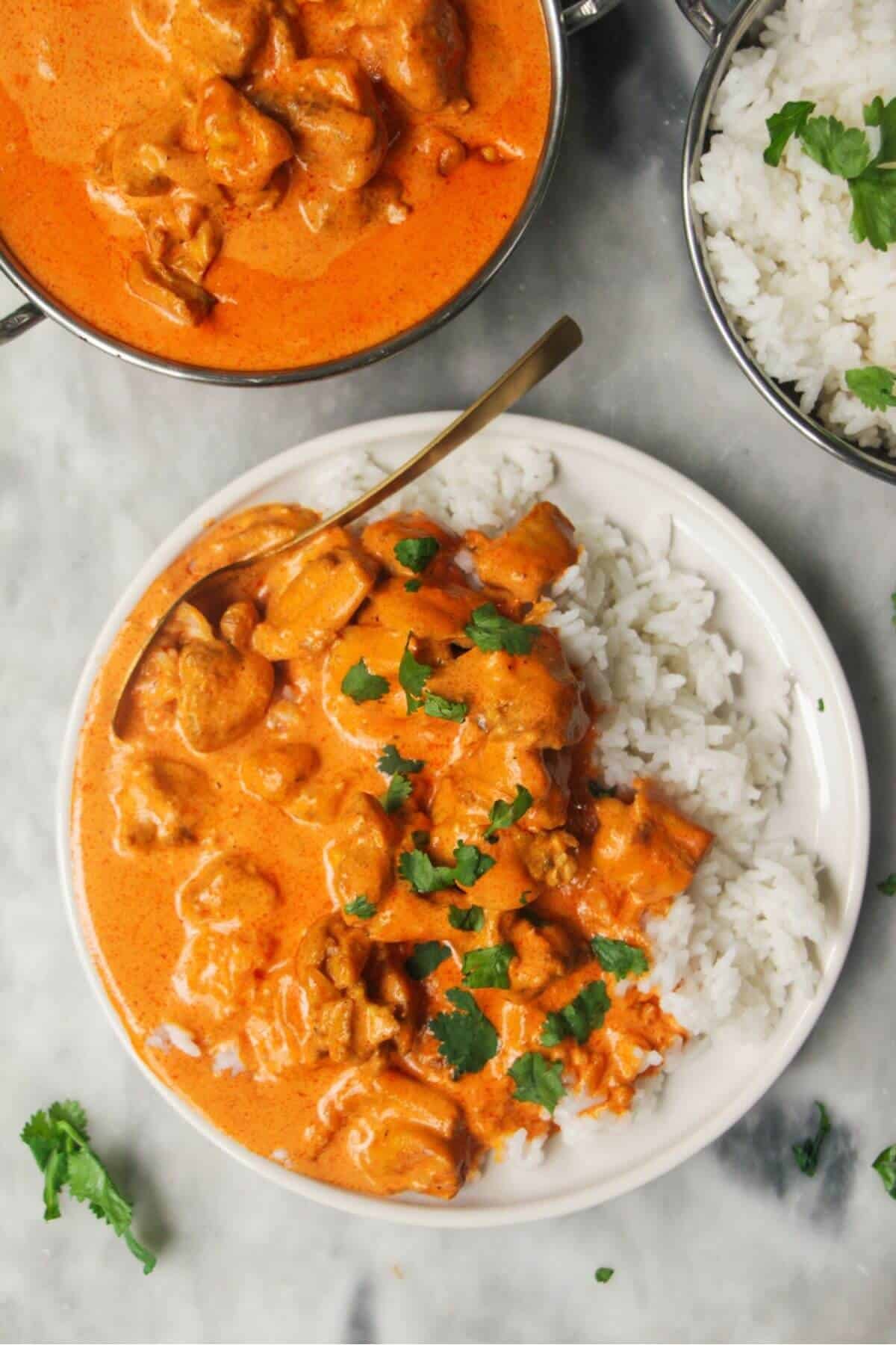 White plate with rice and butter chicken with bowl of butter chicken in the background and garlic naan.
