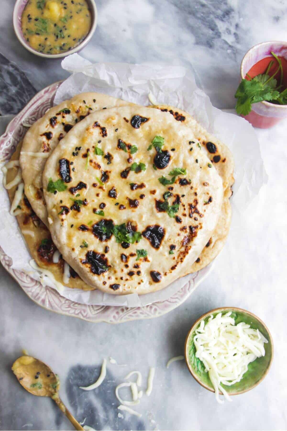 A pile of cheese naan on a small pink plate with a small bowl of grated cheese and butter on the side.