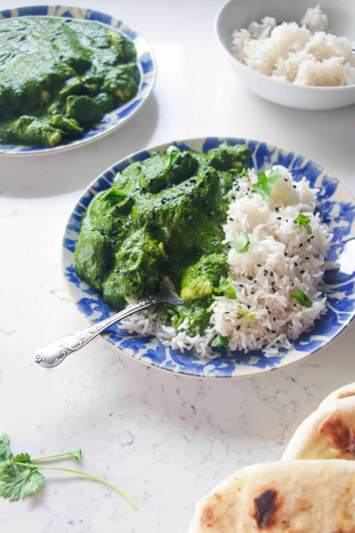 Chicken saag on a white and blue plate with rice, with more curry and naan on the side.
