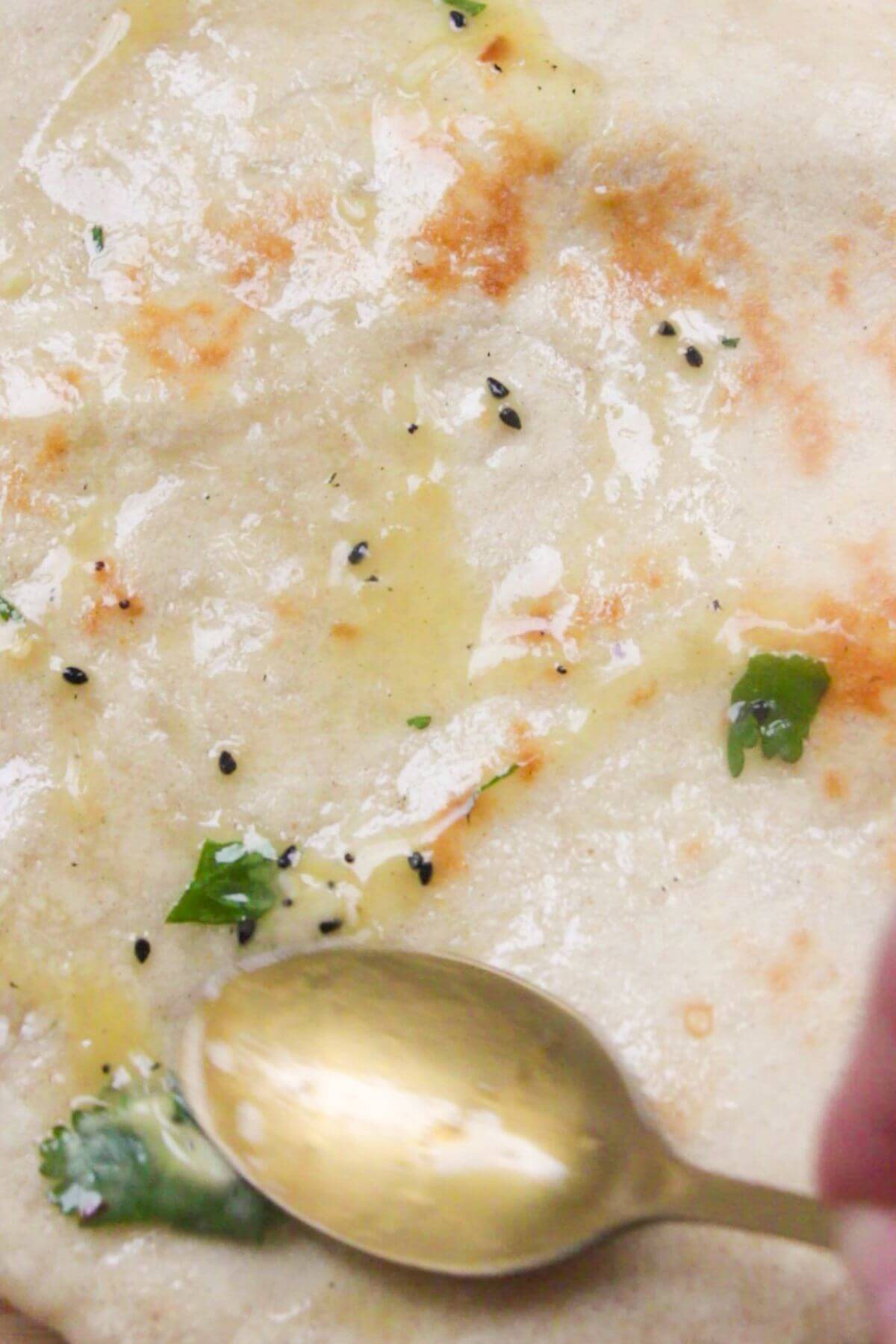 Close up of a small gold spoon smoothing garlic butter onto cooked naan.