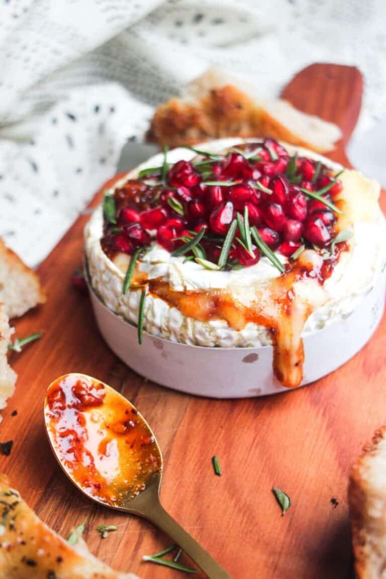 Wheel of camembert on a wooden board with oozy cheese spilling out, topped with pomegranate seeds and herbs.
