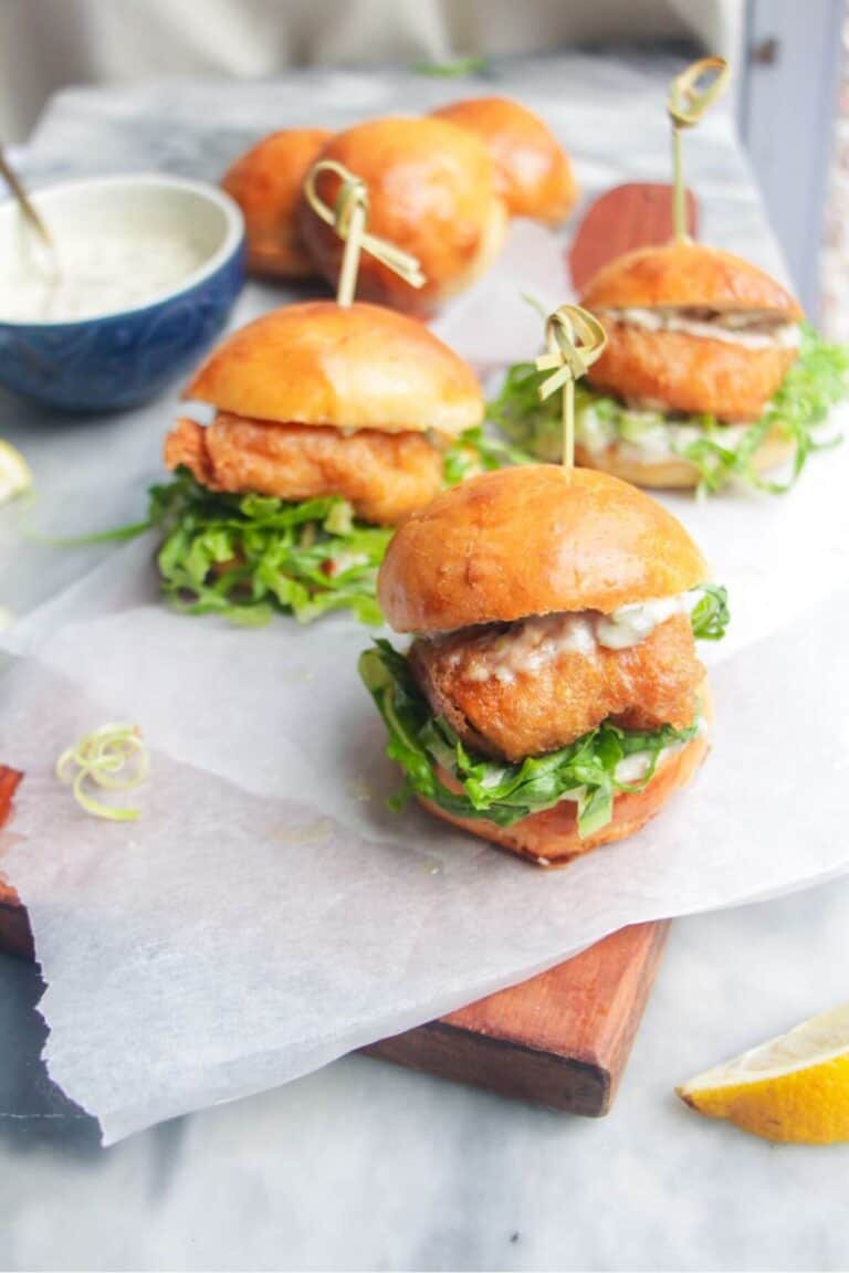 Three beer battered fish sliders on a baking paper lined wooden board with tartare sauce on the side.