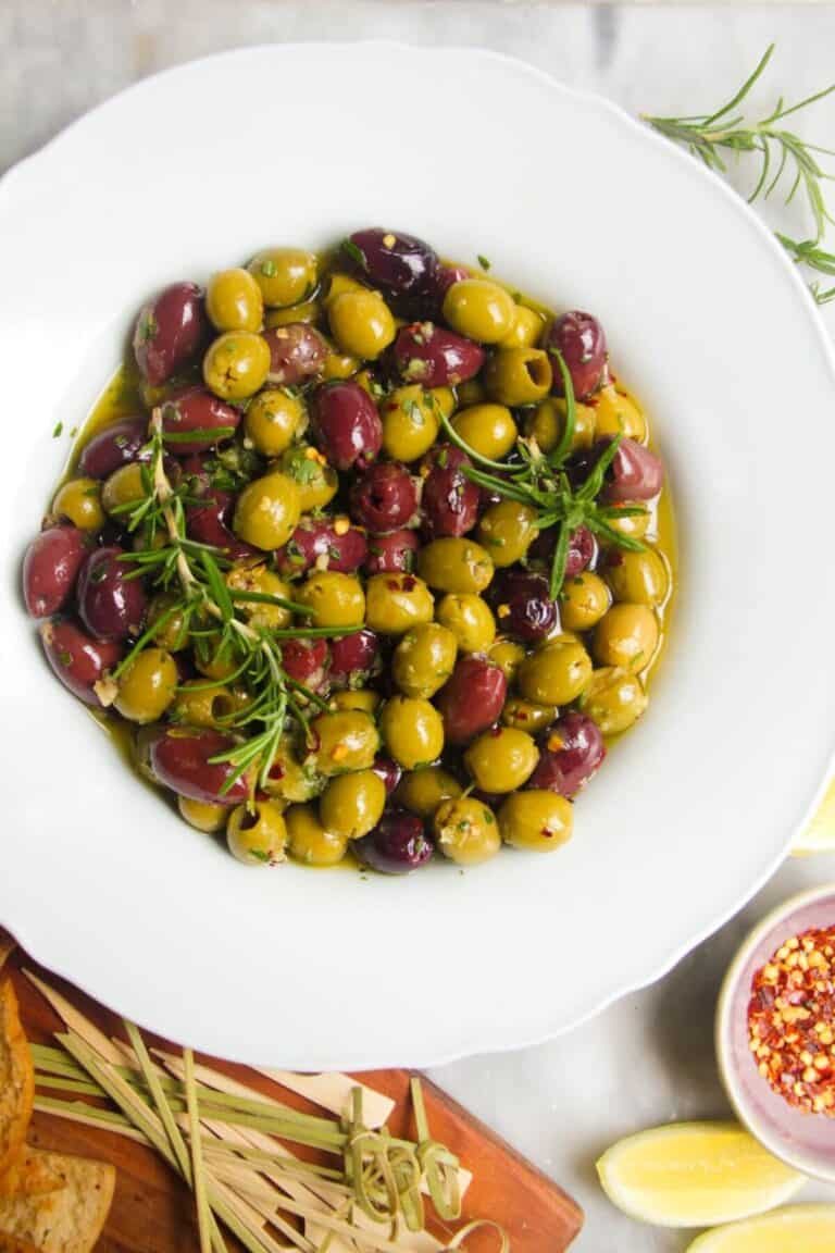 Close up of a white scalloped bowl of marinated olives, with skewers on the side.