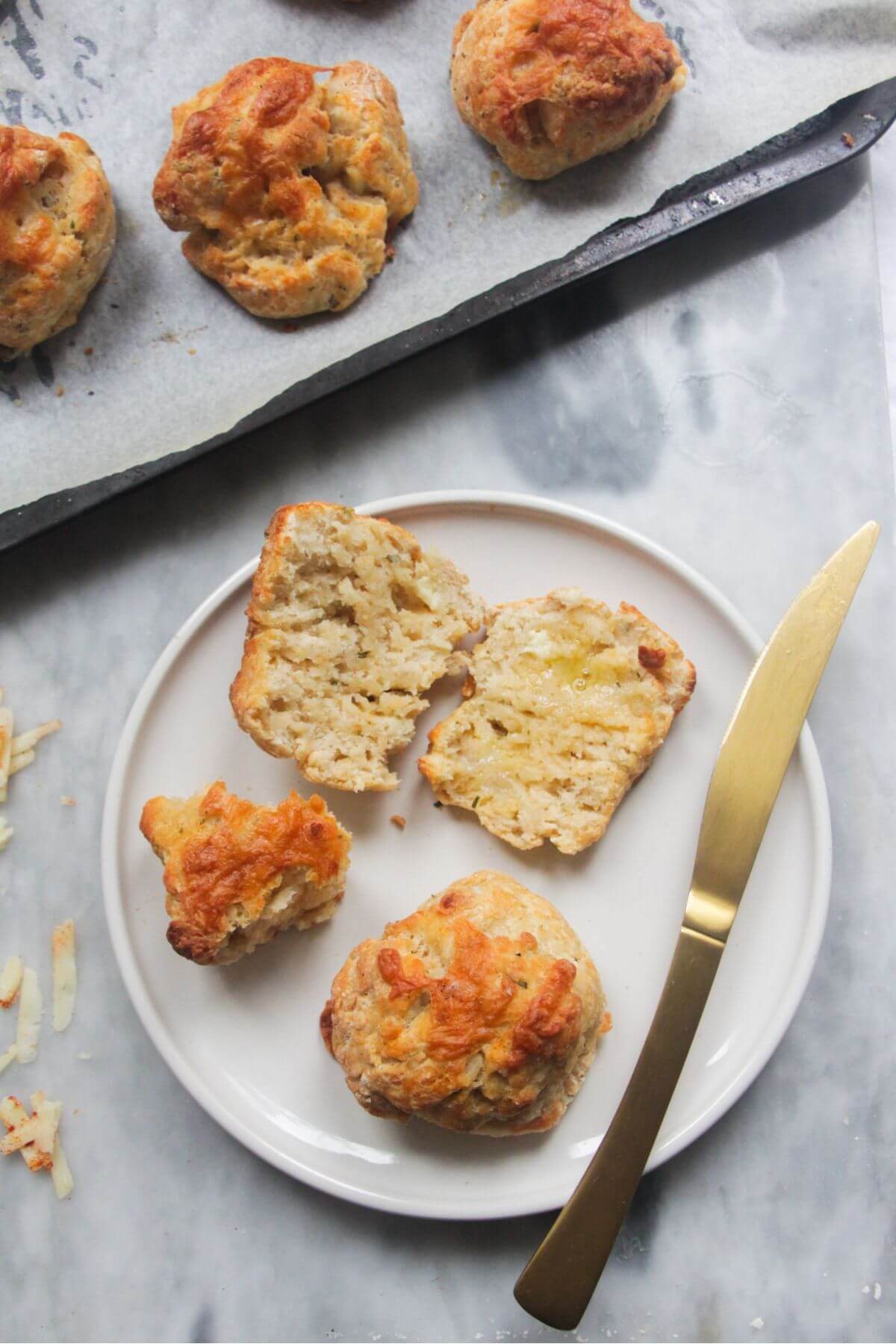 Small white plate with a whole and a sliced open cheese scone, with a gold knife on the side and tray of scones in the background.