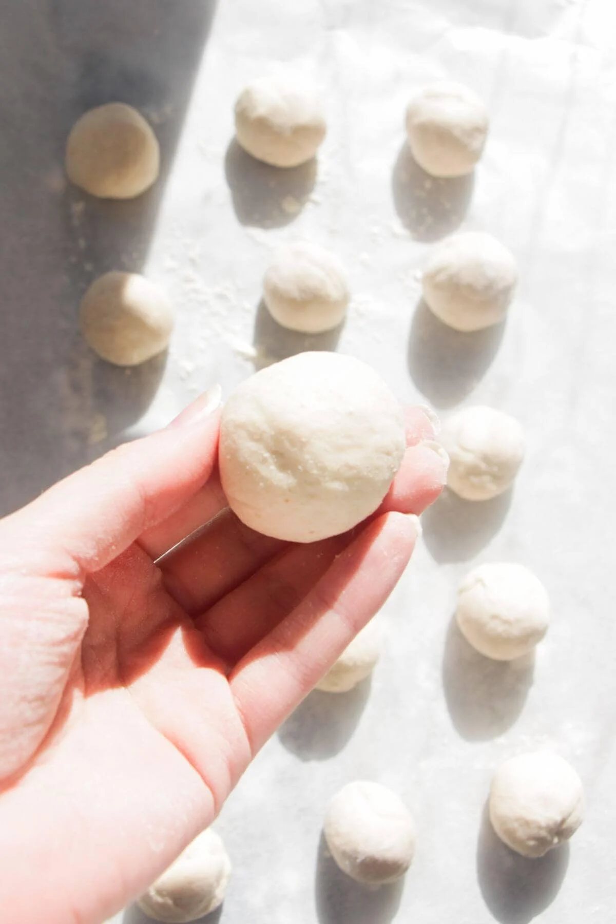 Hand holding up a small ball of dough, with more dough balls in the background.