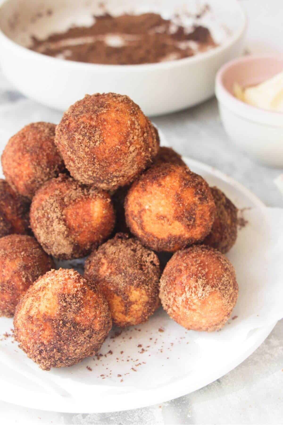 A pile of sugar coated donut bites on a small white plate, with bowl of more pumpkin spice sugar in the background.