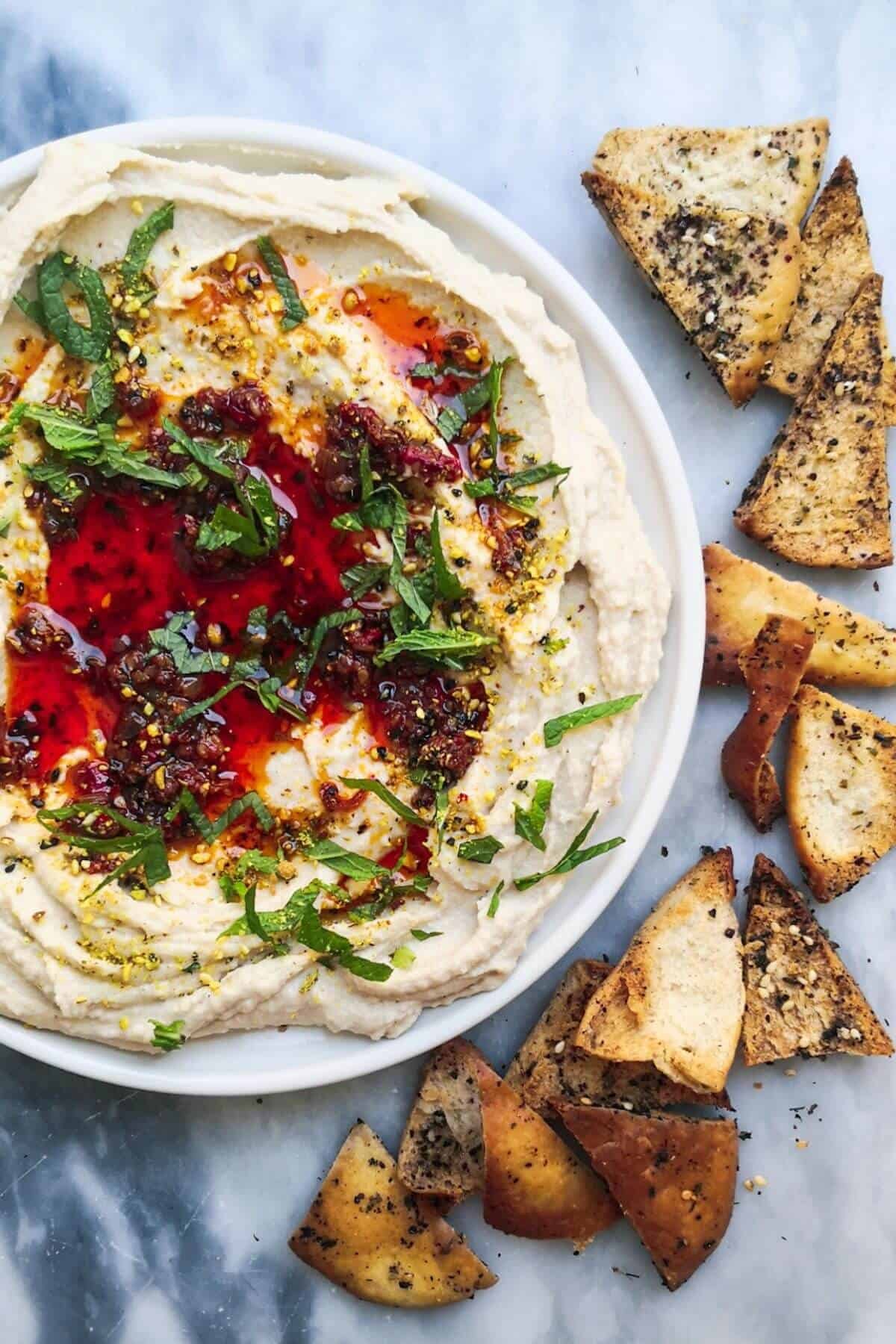 A small white plate of chilli oil topped super smooth hummus, with pita chips on the side.