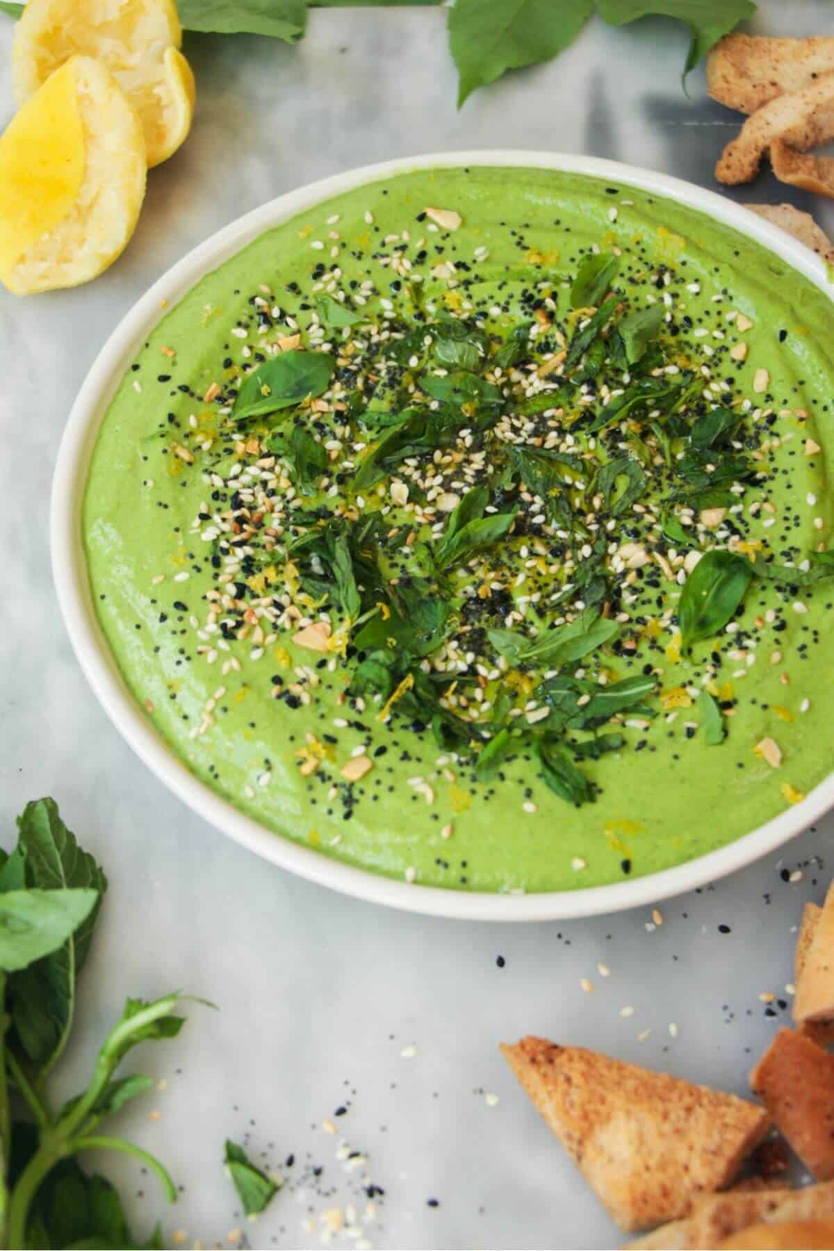 Green goddess dip on a small white plate, with herbs on top and pita chips on the side.