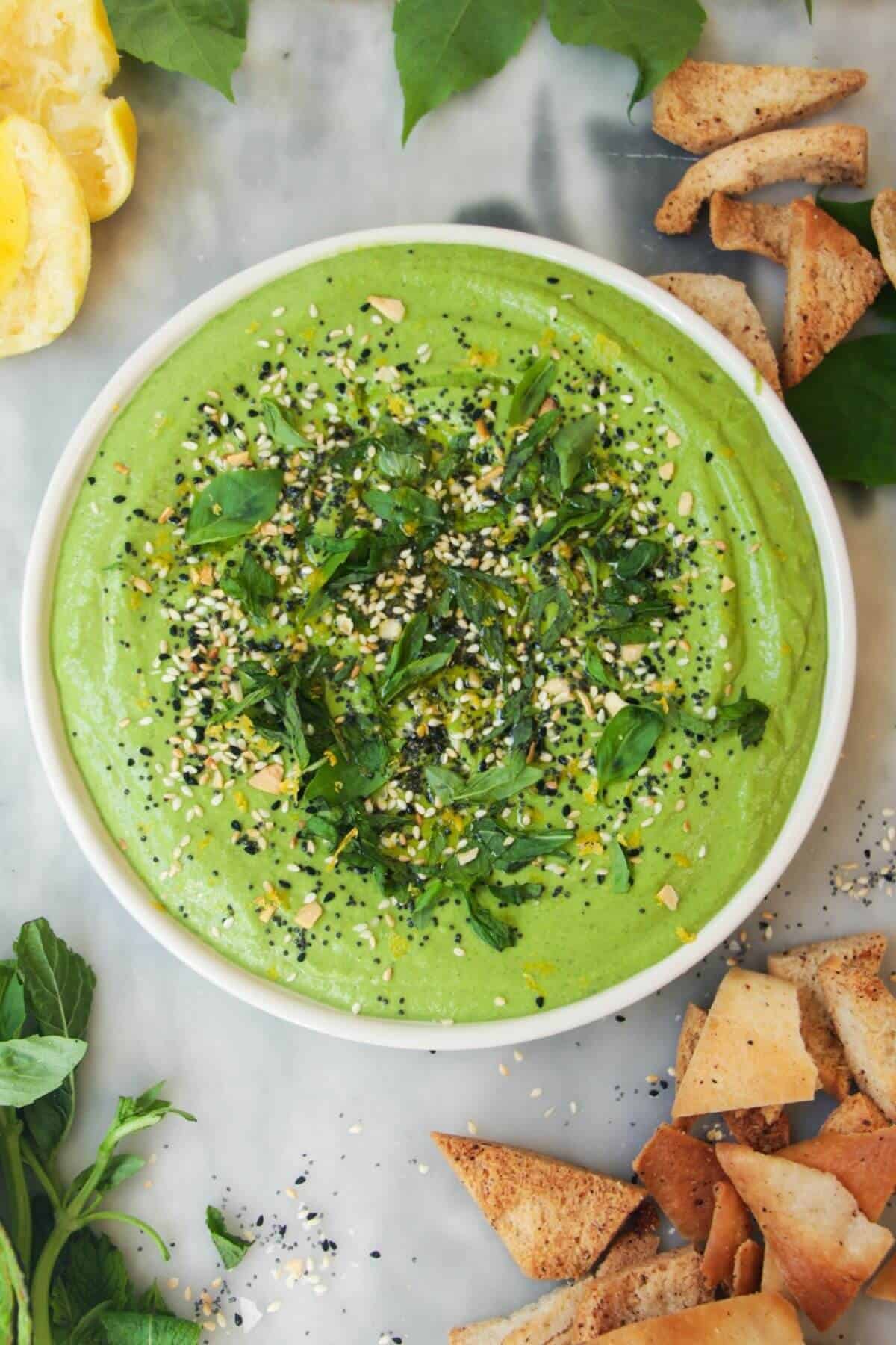 Green goddess dip on a small white plate, with herbs on top and pita chips on the side.
