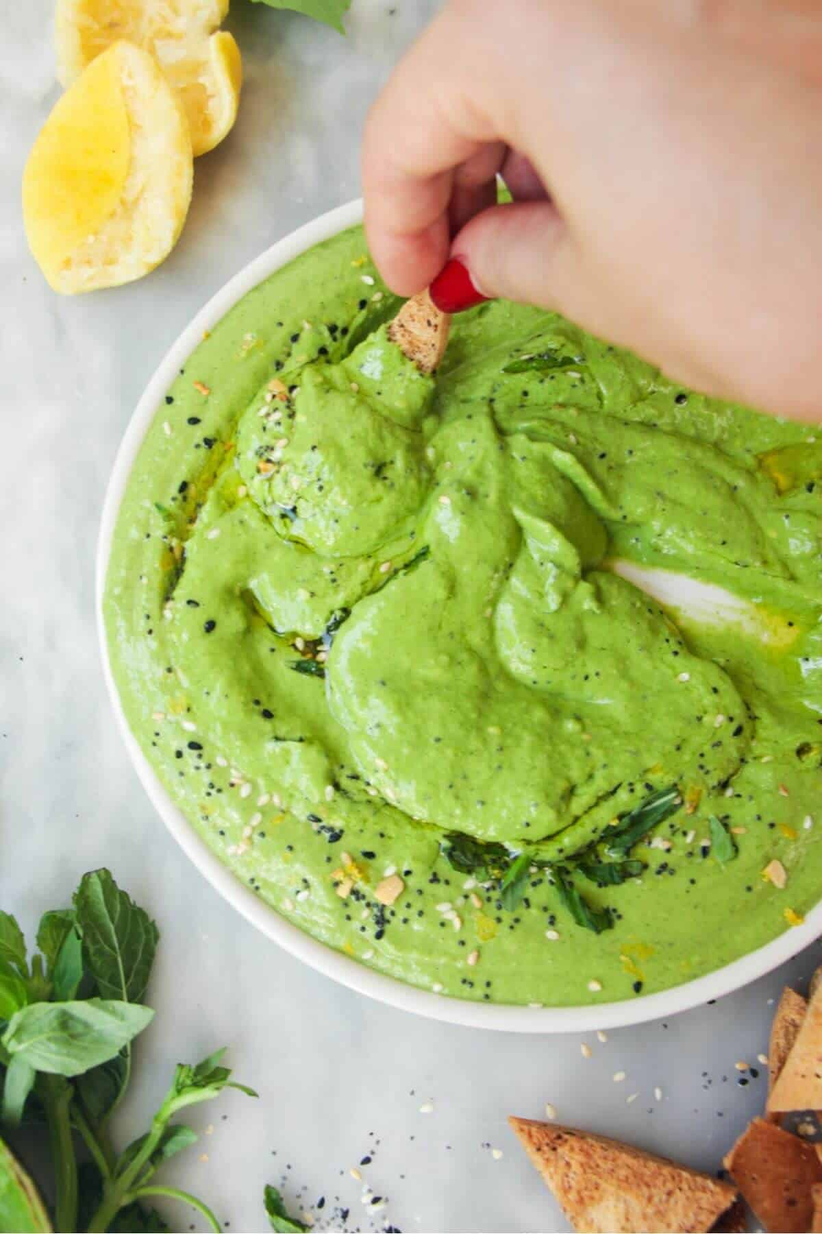 Hand swirling a pita chip through green goddess dip on a white plate.