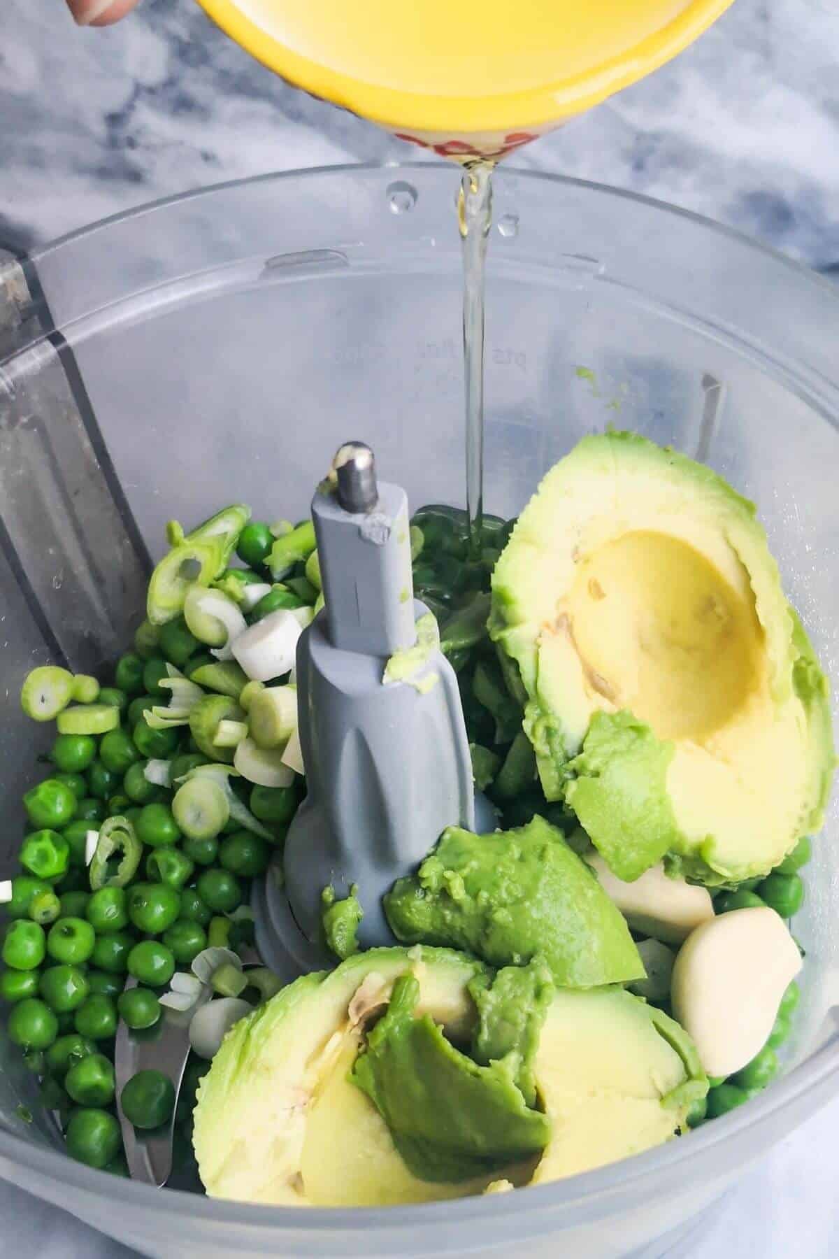 Olive oil being added to the bowl of a food processor with avocado, garlic and peas.