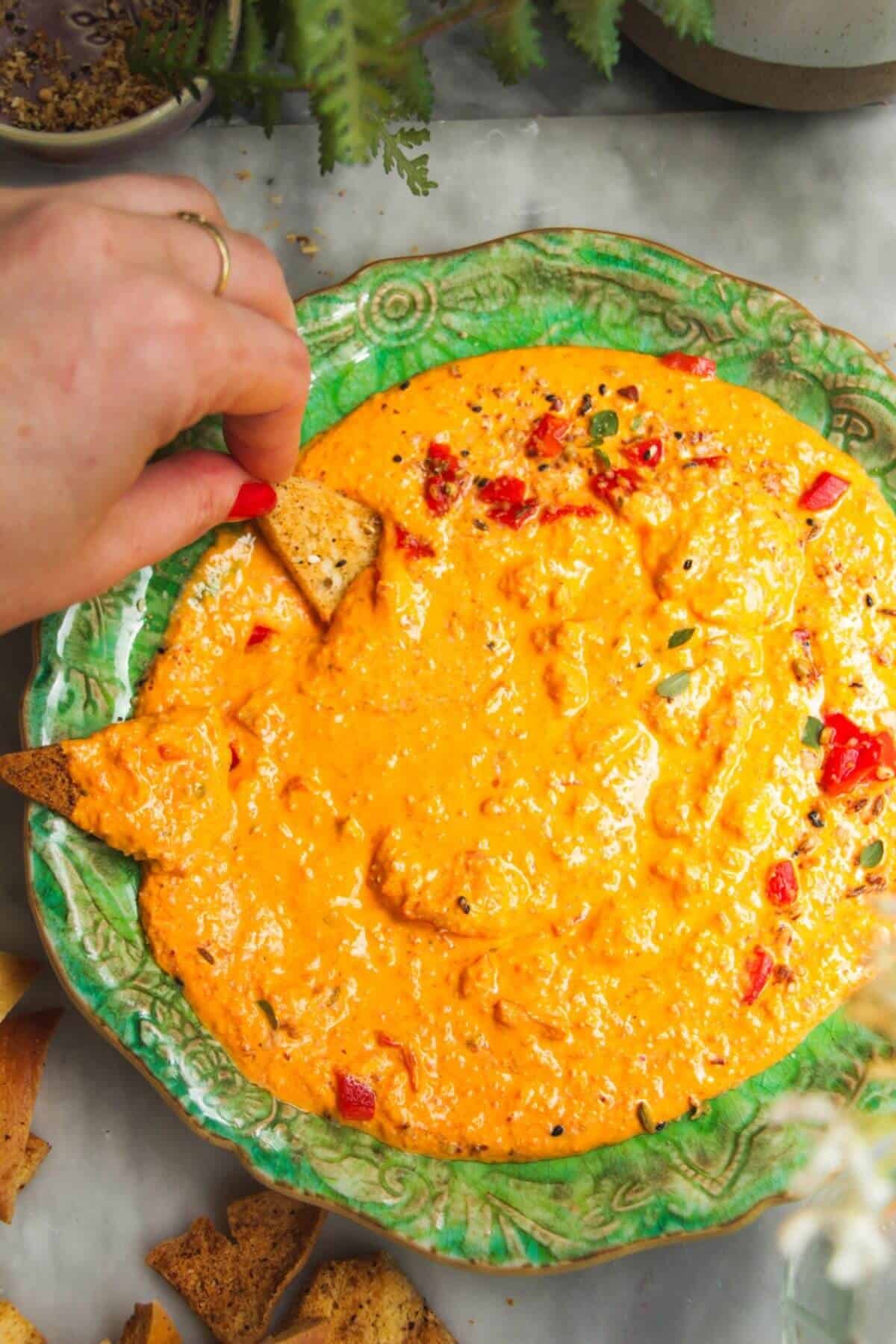 Hand scooping pita chip through red pepper dip.