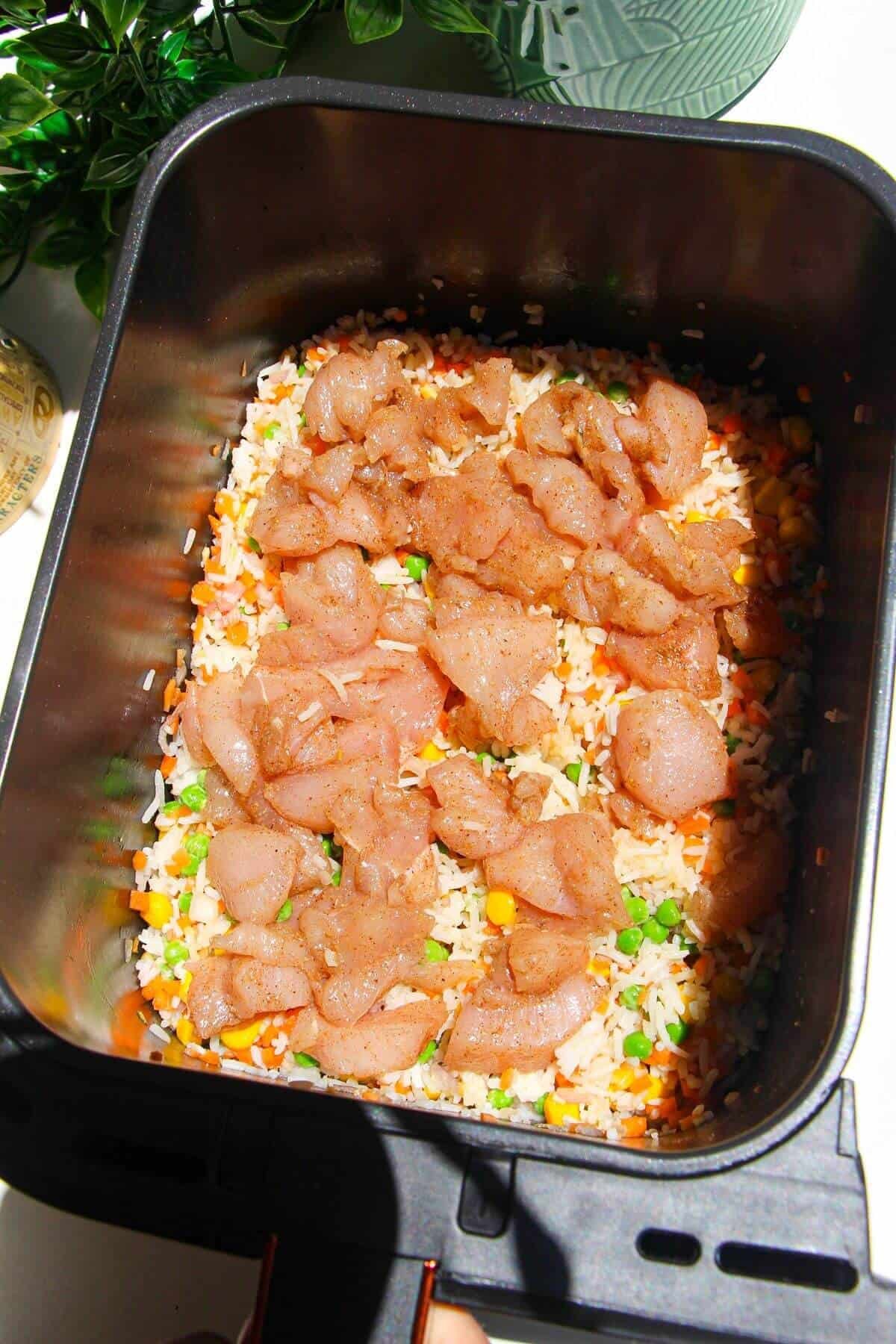Seasoned chicken on top of rice and vegetables in air fryer basket.