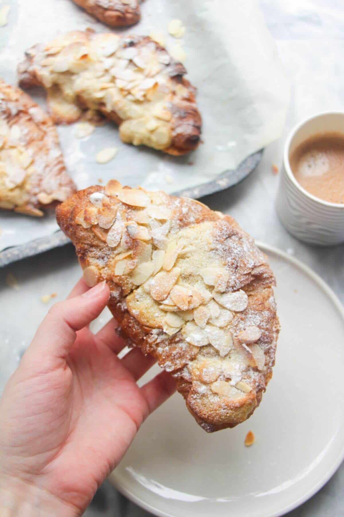 A hand holding an almond croissant with more almond croissants in the background.