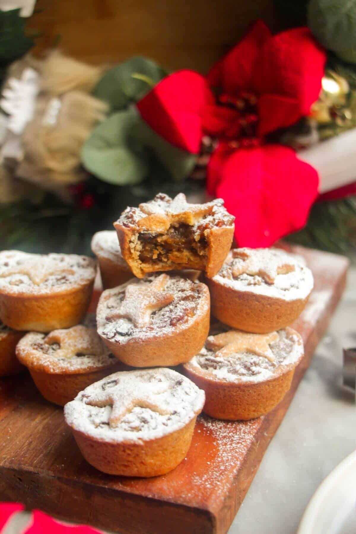 A pile of icing sugar dusted Christmas mince pies on a small wooden board with Christmas deocrations in the background.
