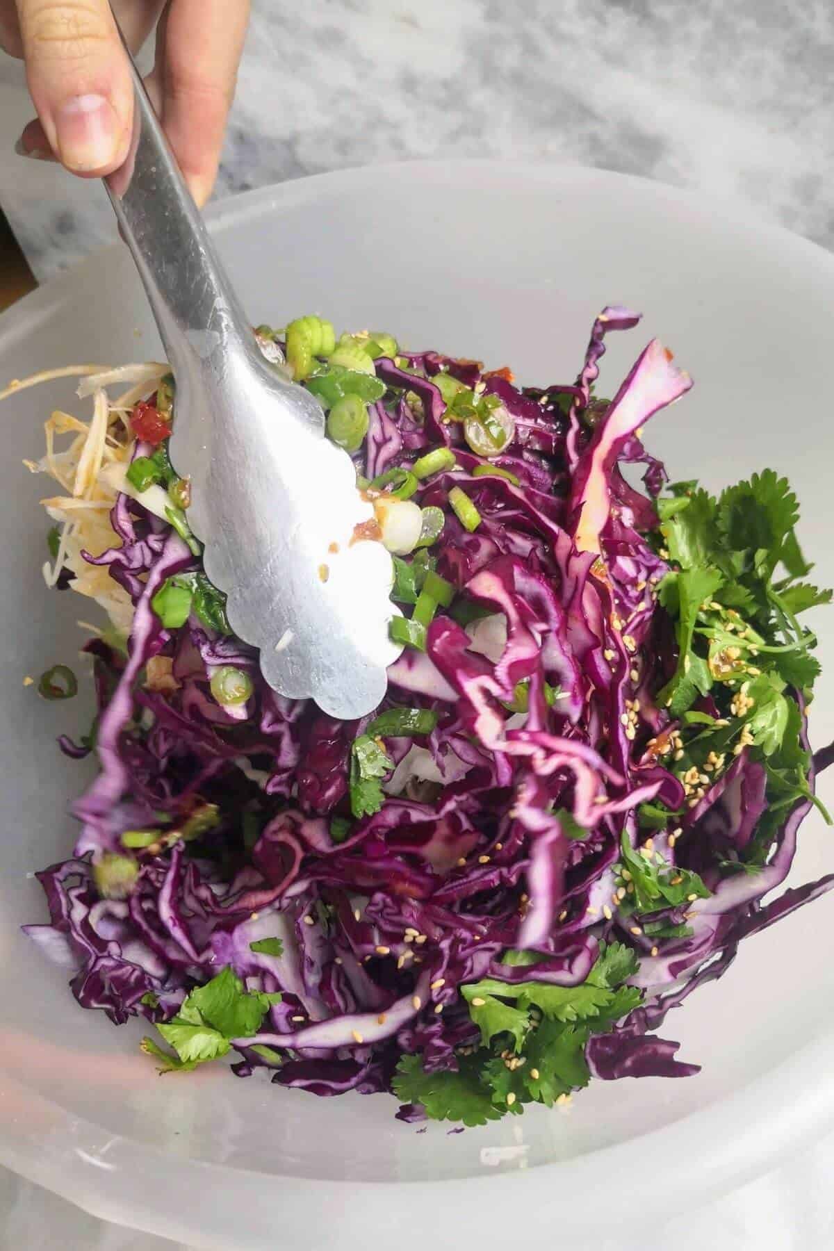 Asian slaw mixed with dressing with silver tongs.