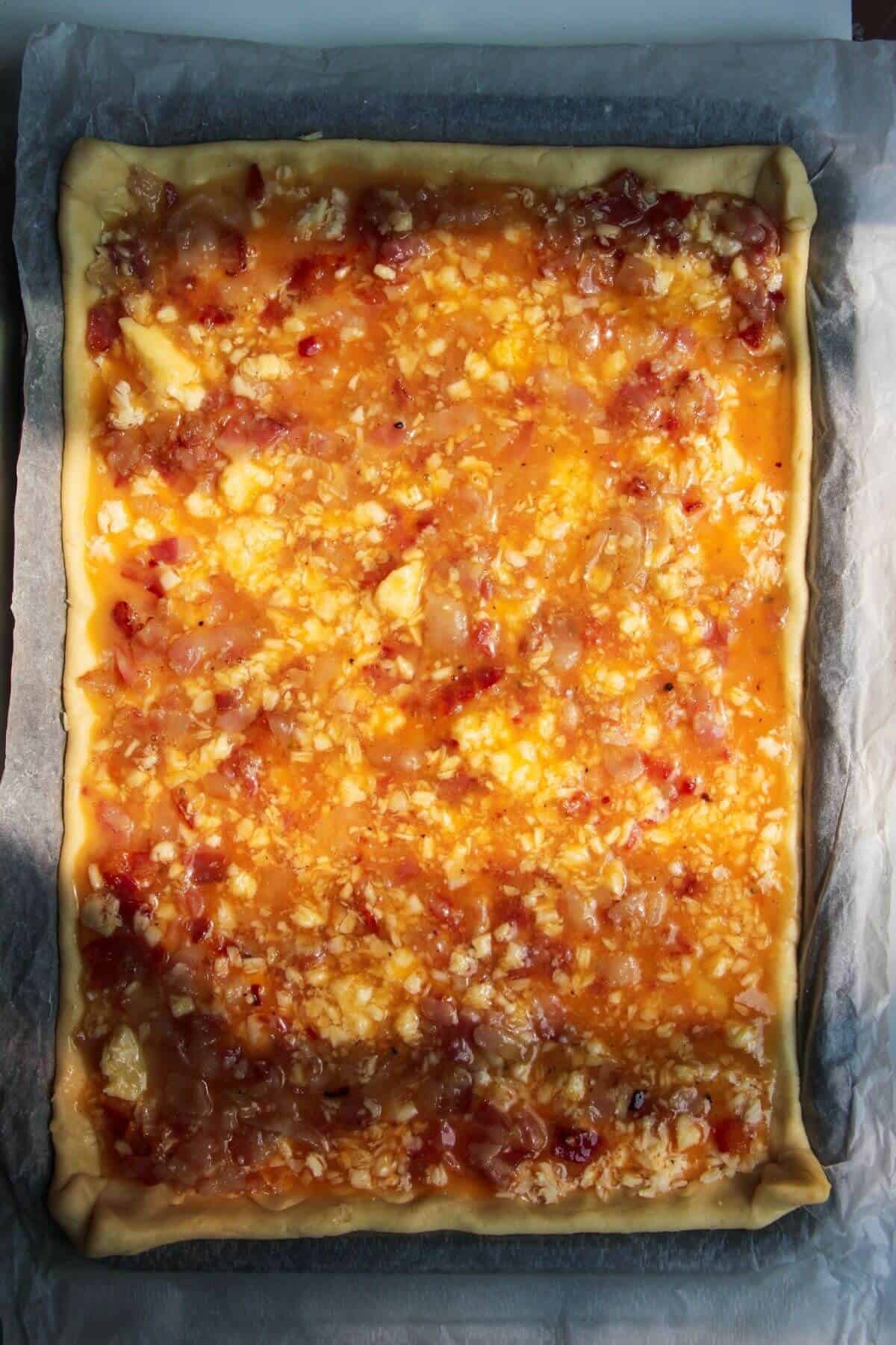 Whisked egg poured on top of bacon and cheese in pastry, on a lined oven tray.