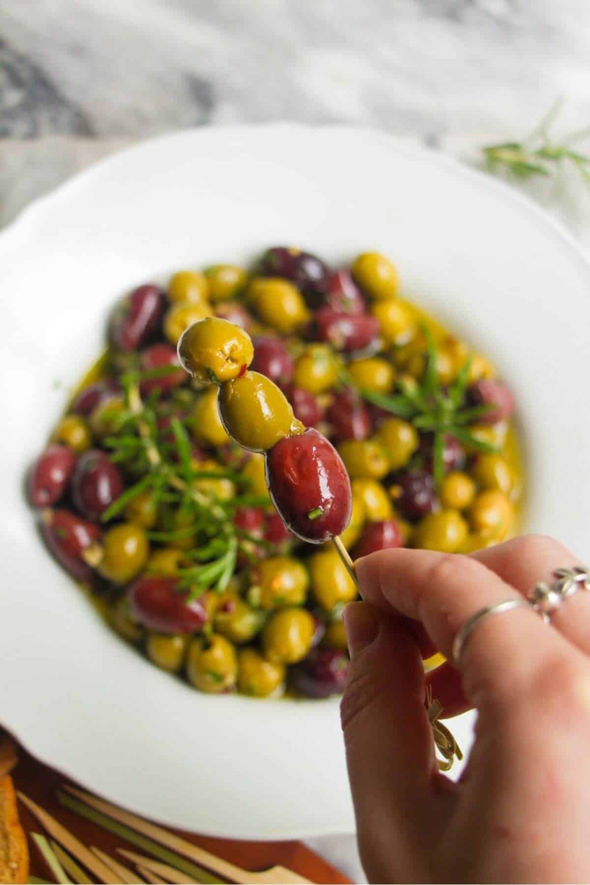 A hand holding up three olives on a skewer with a bowl of marinated olives in the background.