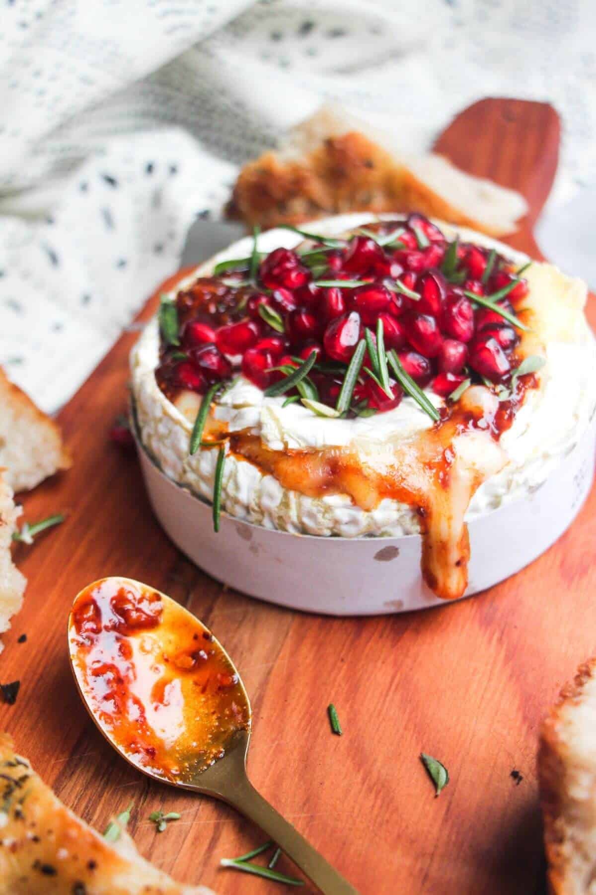 Wheel of camembert on a wooden board with oozy cheese spilling out, topped with pomegranate seeds and herbs.
