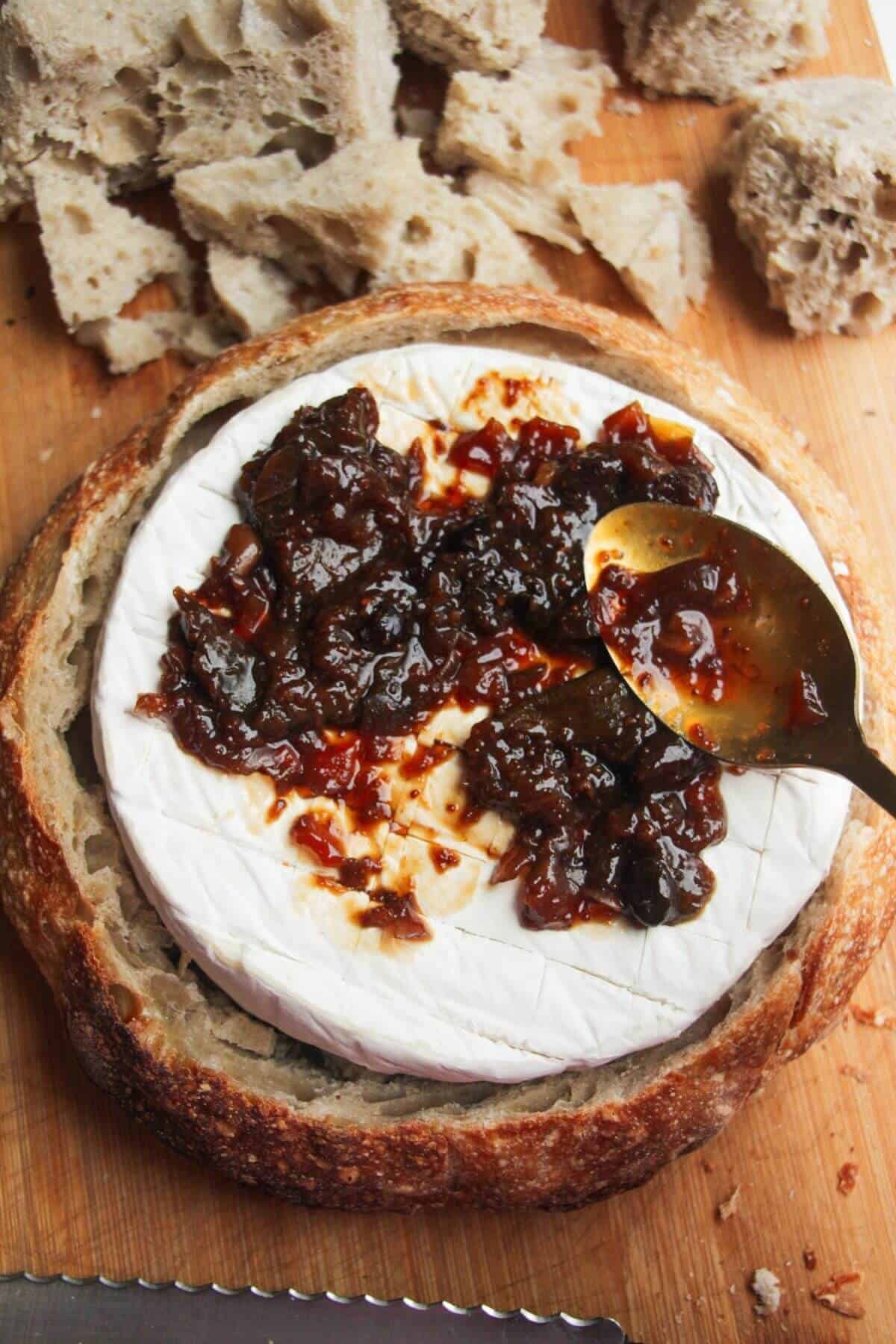 Spooning fig jam on top of brie in a bread bowl.