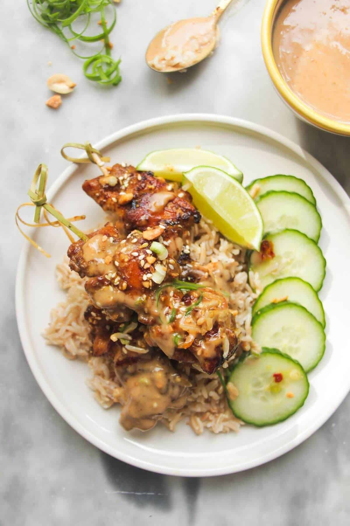 Chicken satay skewers on rice with cucumber slices on the side, with a small bowl of satay peanut sauce behind.