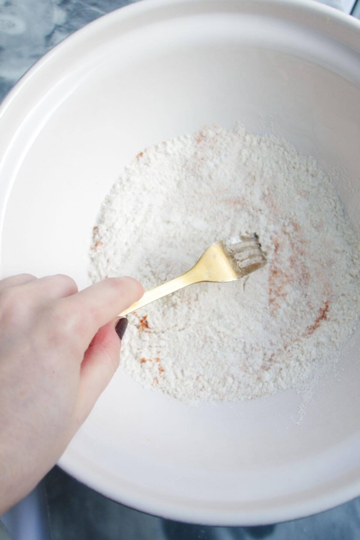 Hand holding a gold fork whisking flour and turmeric in a large white mixing bowl.