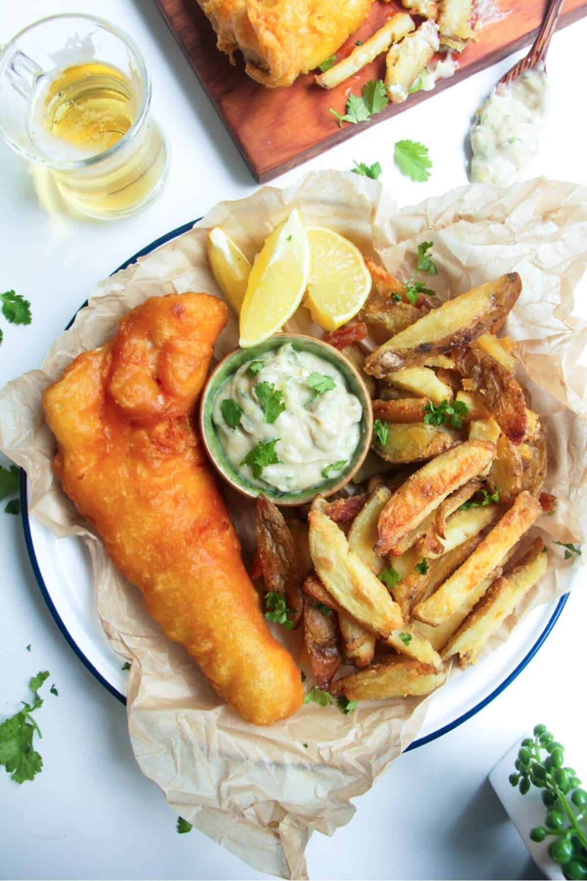Beer battered fish fillet with a pile of chips and a small pot of tartar sauce in baking paper lined plate.