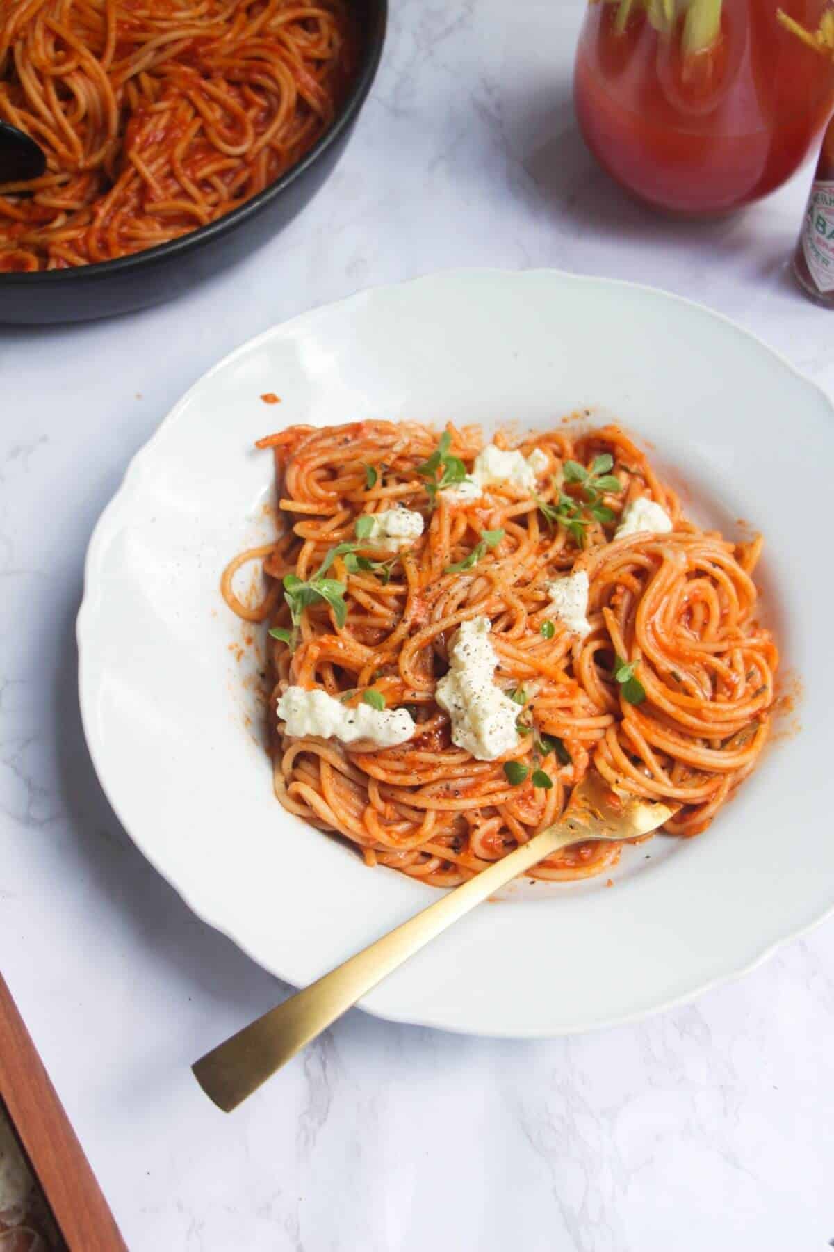 Bloody mary pasta, topped with burrata and basil, in a white scalloped bowl with more pasta in the background.