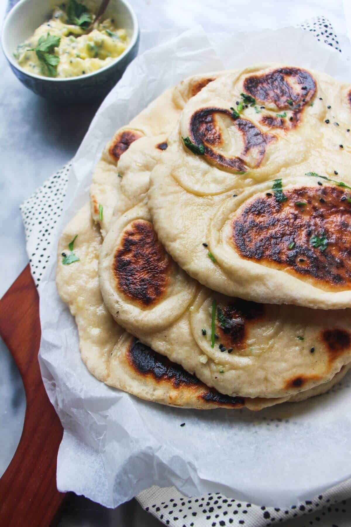 A pile of garlic naan with charred spots on baking paper on a wooden board, with a small bowl of garlic butter in the background.