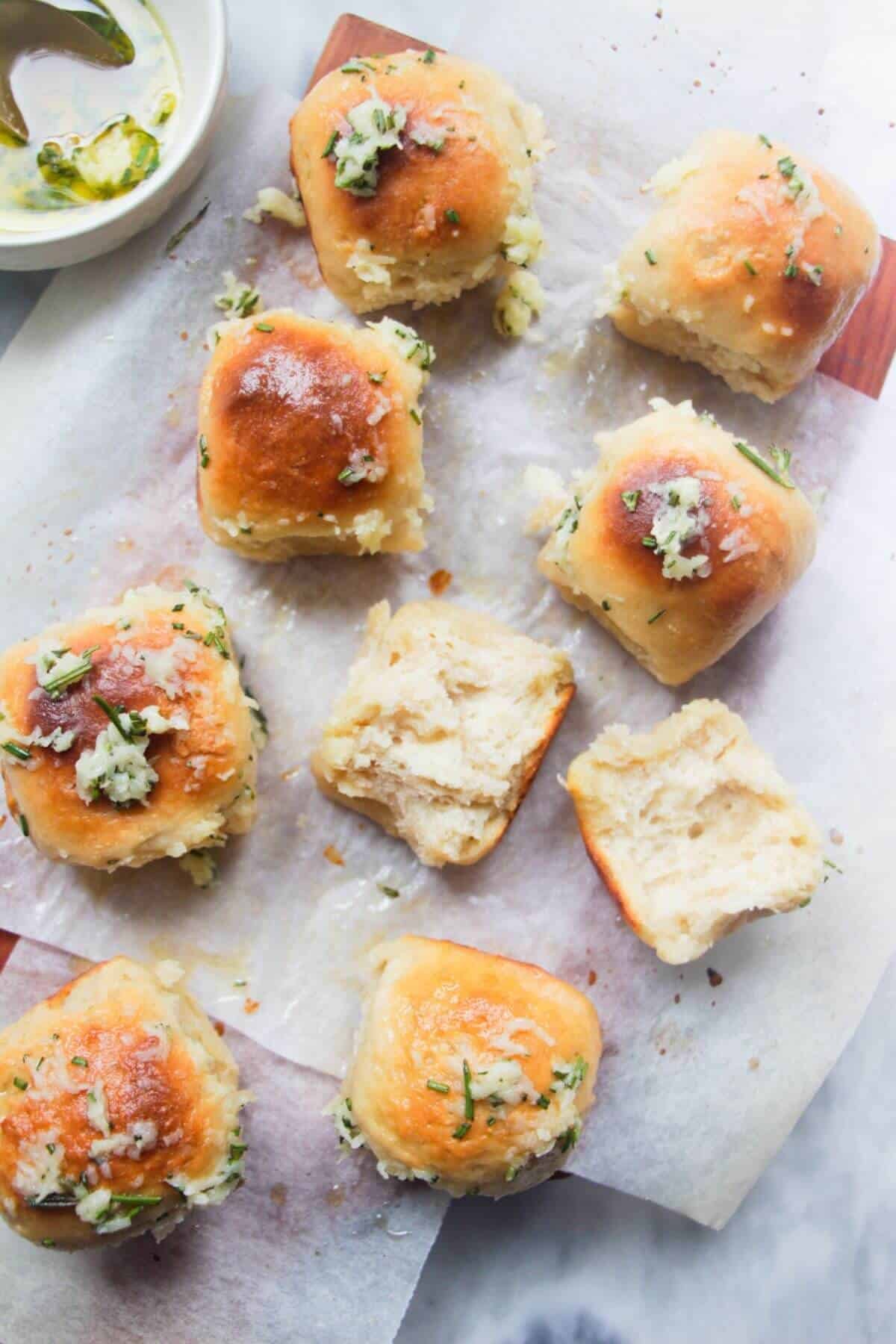 8 garlic butter topped rolled assembled on a baking paper lined wooden board with bowl of garlic butter in the background and one roll torn open.