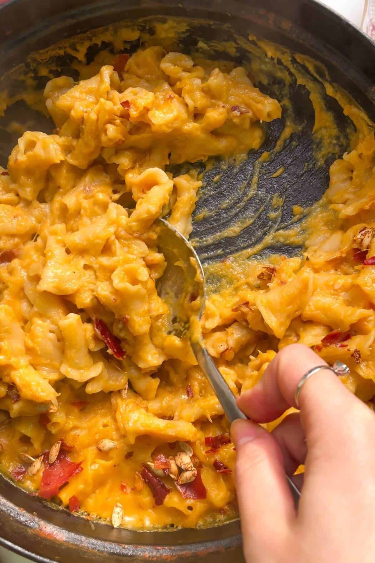 Close up of hand scooping up butternut squash mac and cheese with a large spoon from the pan.