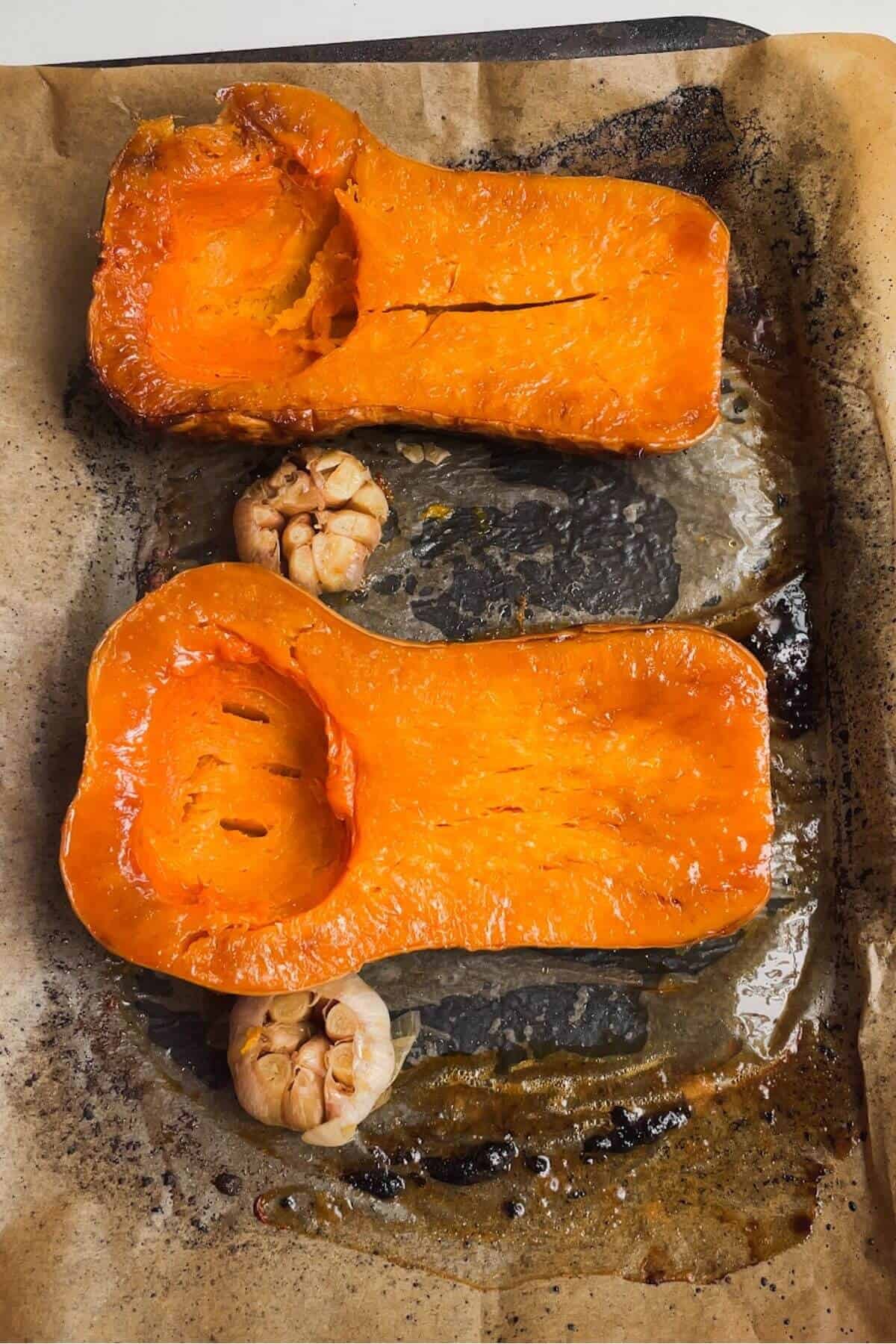 Two halves of a roasted squash on an oven tray, with two heads of garlic.