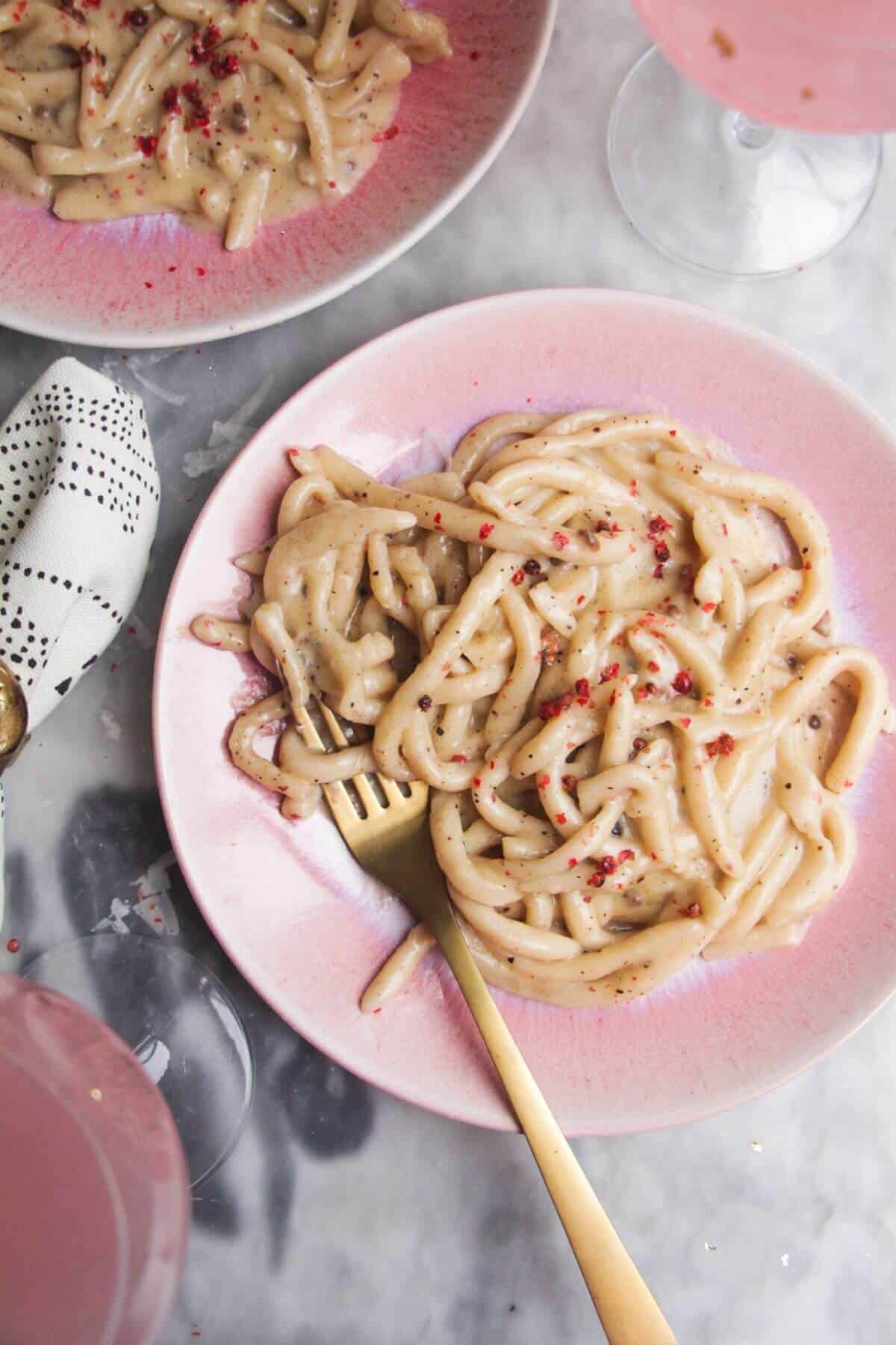 Silky, cheesy cacio e pepe on a pink plate, with a gold fork and another plate in the background.