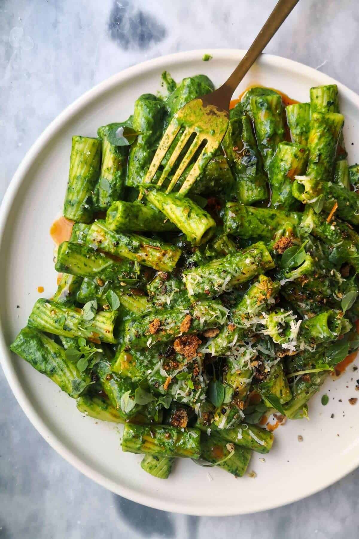 Green pasta sauce with rigatoni on a white plate, with gold fork piercing a piece of pasta.
