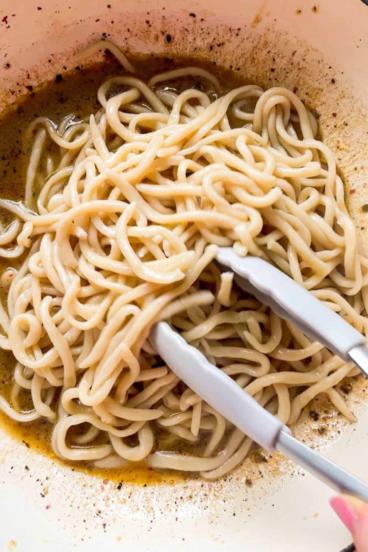 Tongs adding cooked pasta to butter and black pepper in a pan.
