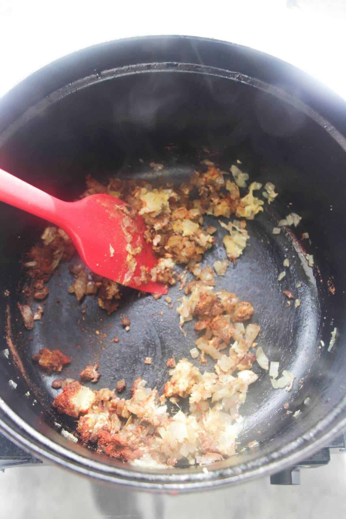 Garlic, ginger, onion and spices being stirred in a large black pot with a large red spatula.