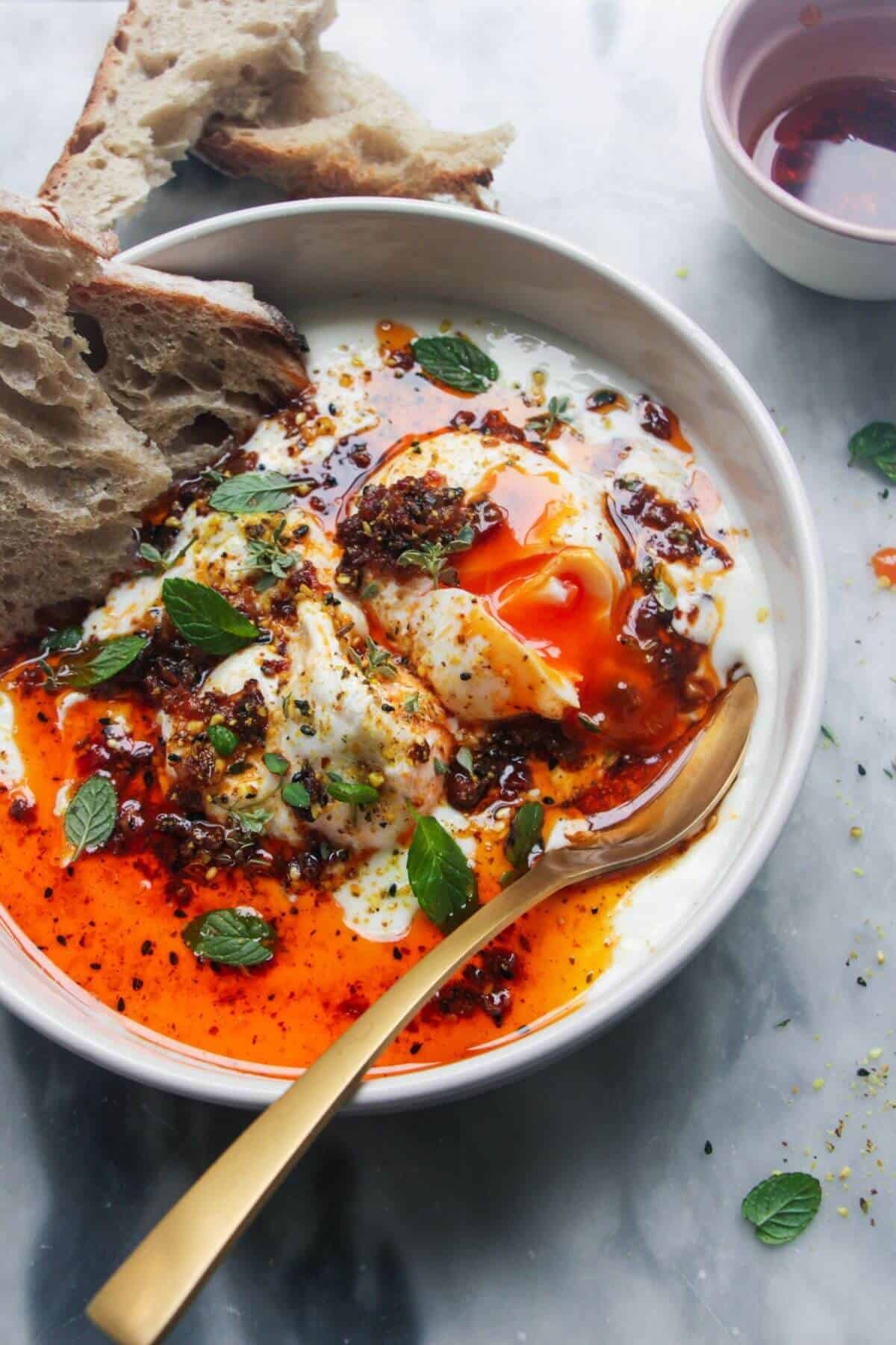 Small white bowl with 2 poached eggs, 1 broken open with yolk coming out, with a gold spoon on the side and toast in the bowl.