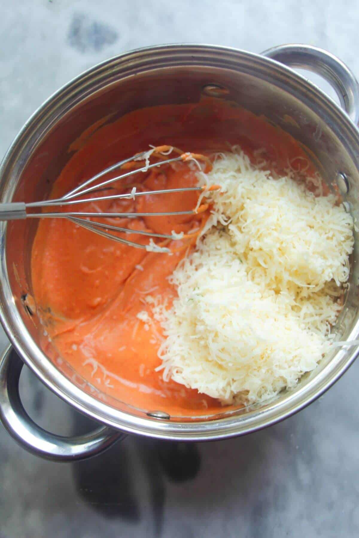 Grated cheese in a small silver pot with gochujang bechamel and a whisk inside.