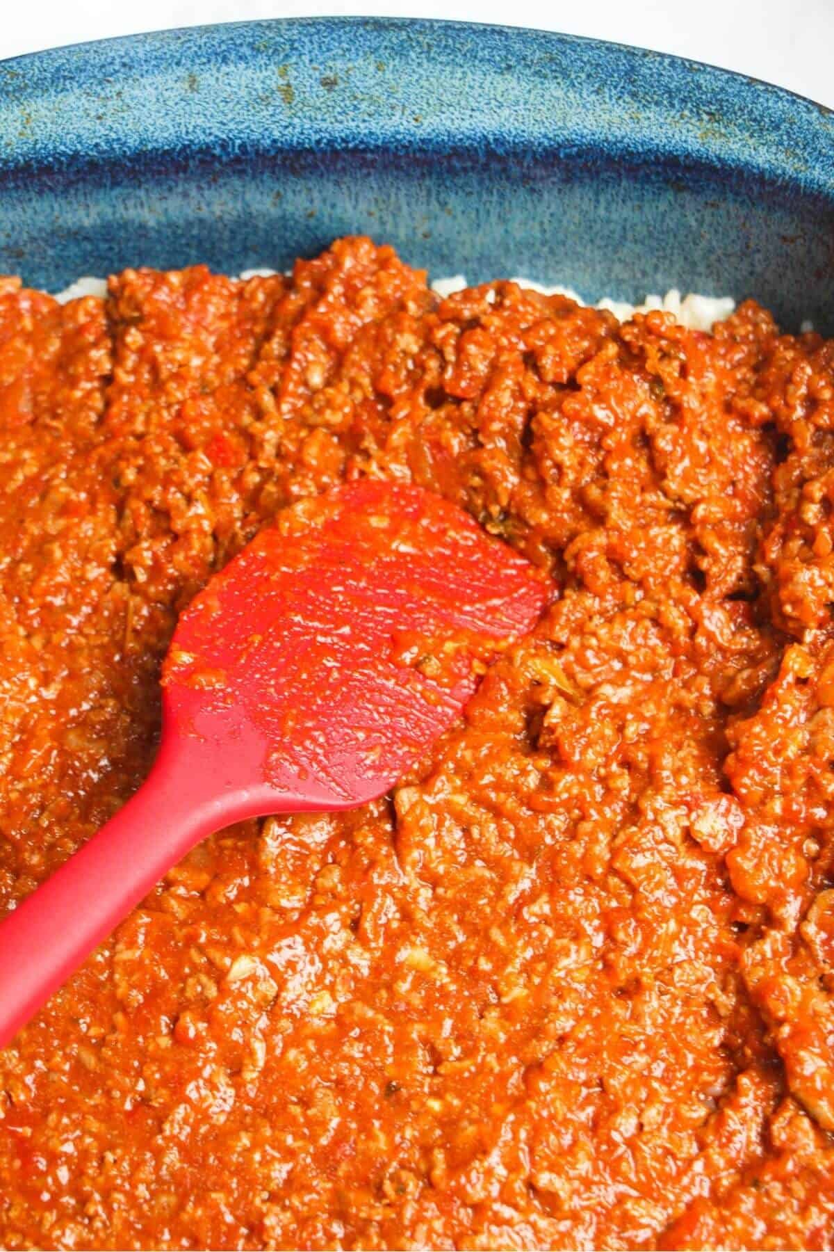Red spatula pressing bolognese into a blue oven dish.