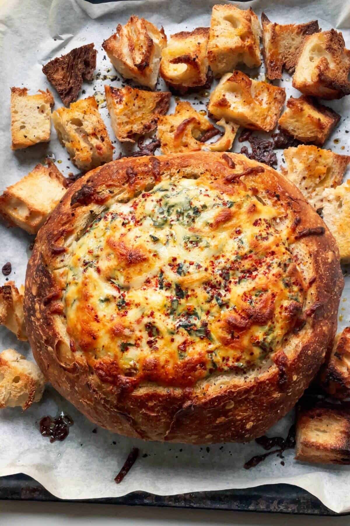 Golden baked dip in a bread bowl on a lined tray, with croutons on the side.