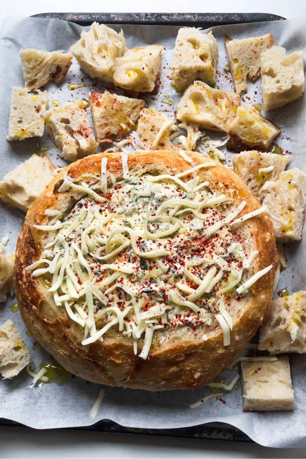 Spinach dip in bread bowl, topped with mozzarella, on a lined tray.