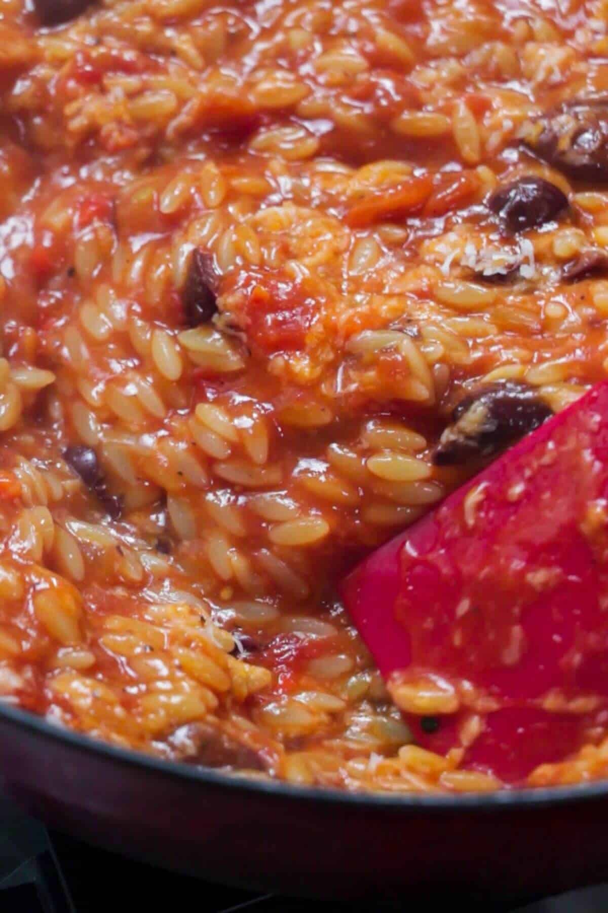 Close up of red spatula stirring tomato orzo in a large black skillet.