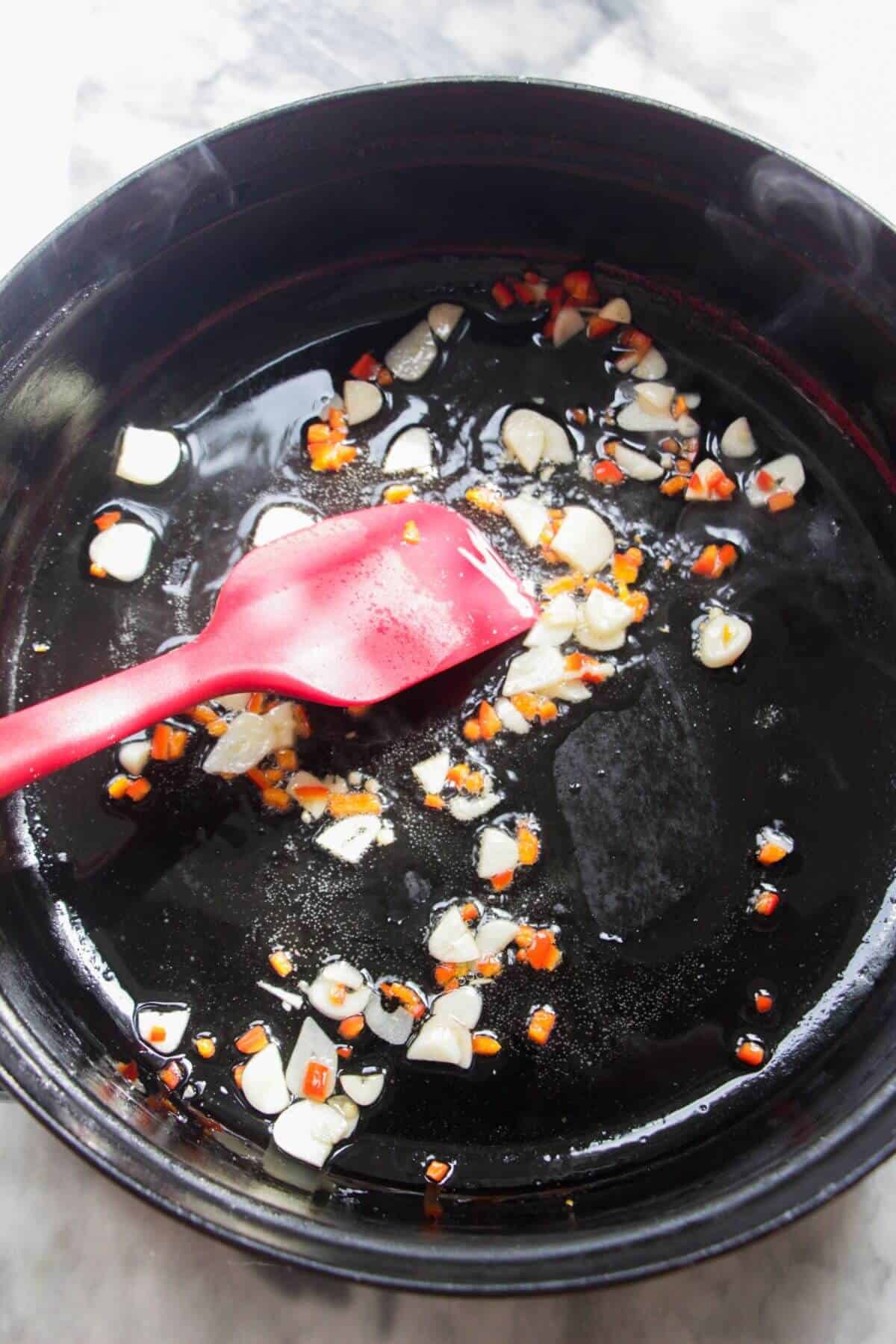 Sliced garlic and chilli being stirred through oil in a large black skillet.